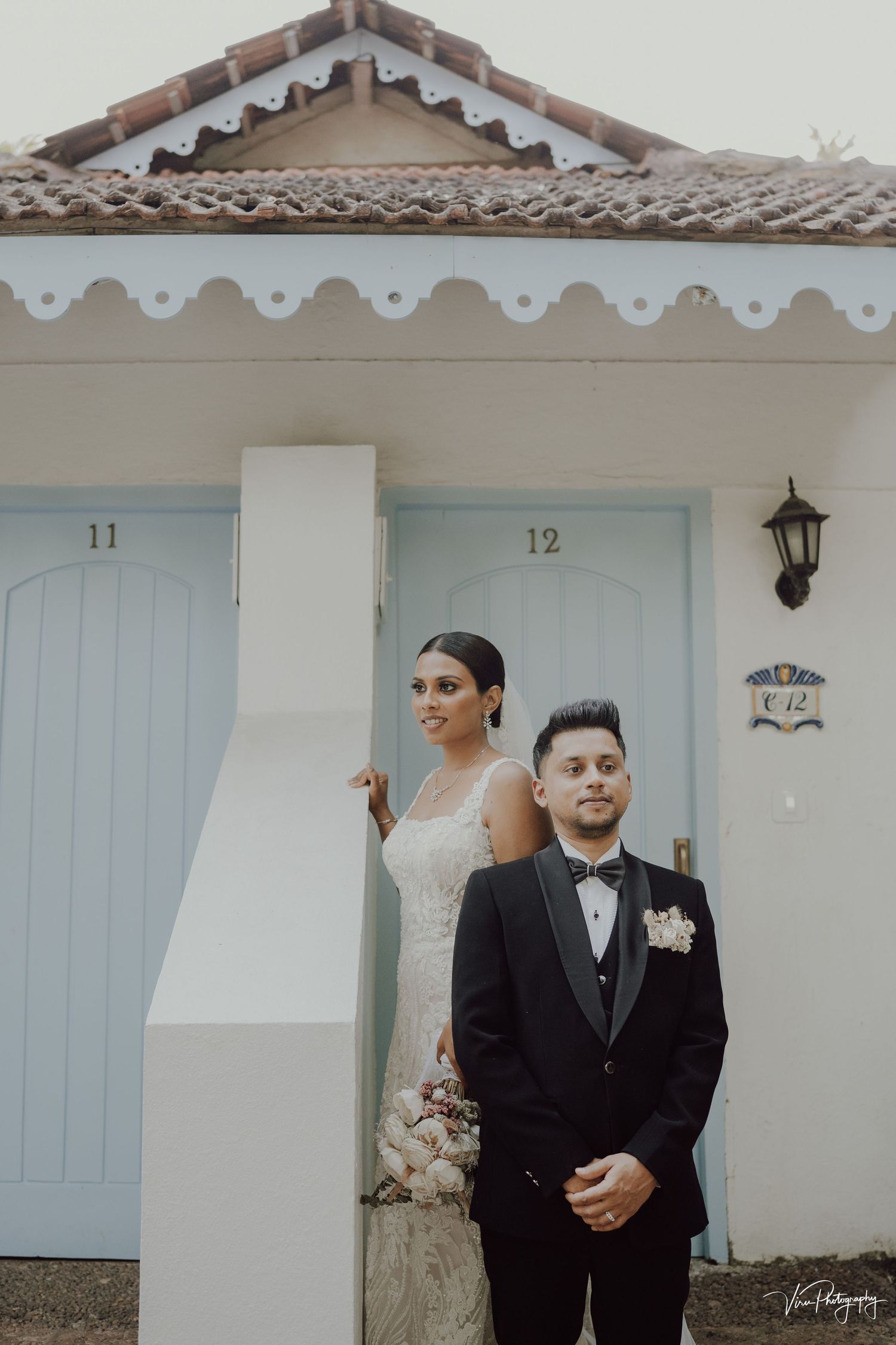A couple stands outside a rustic building with blue doors numbered 11 and 12, featuring decorative white trim and lantern.