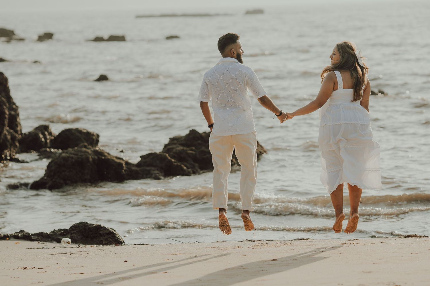 Couple in white outfits walking along a rocky beach shoreline at sunset while waves crash against the coast.