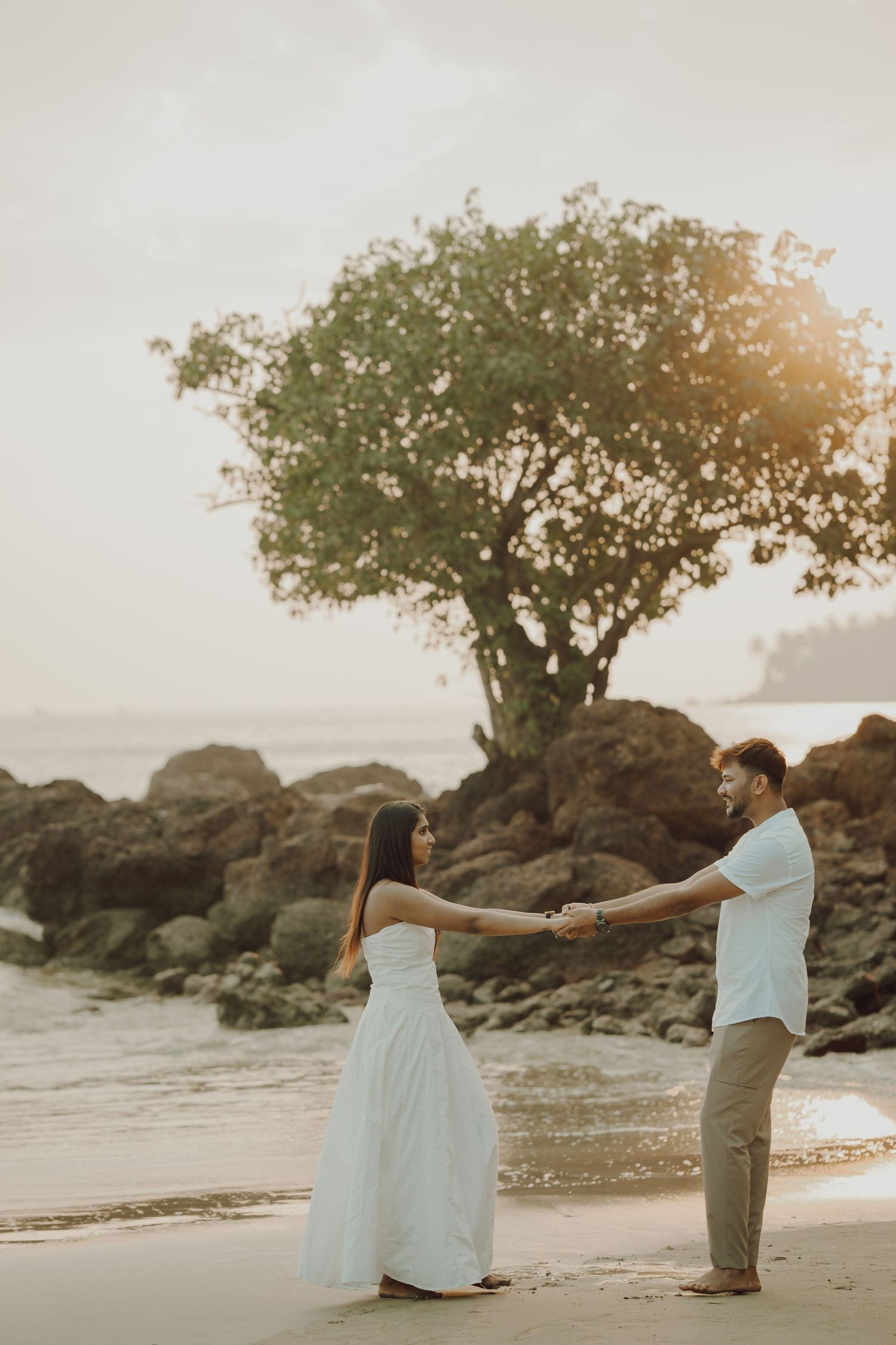 A romantic beach scene at sunset with a couple posing by rocks and trees along a sandy shoreline.