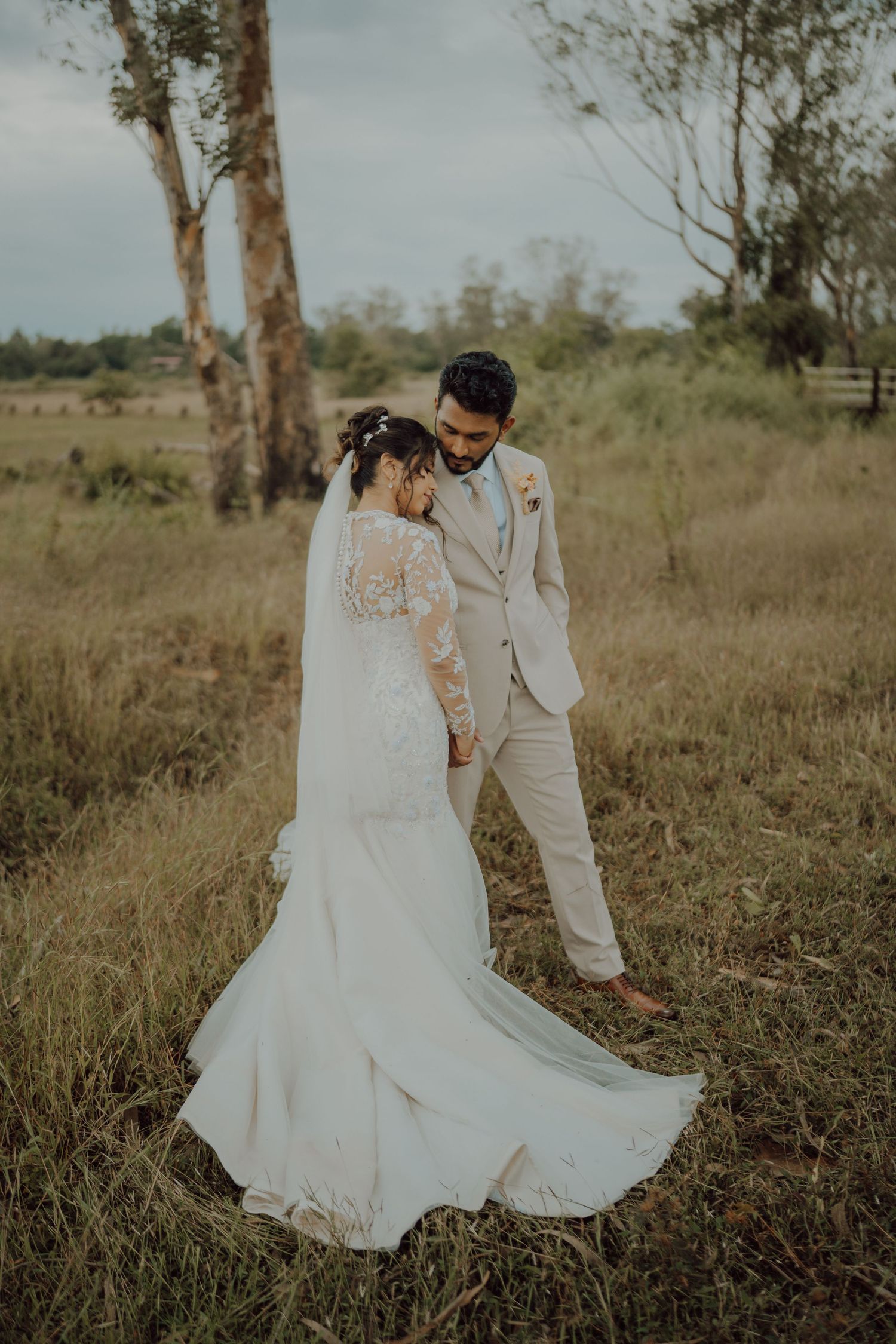 A groom in a beige suit leans toward his bride in a flowing lace gown and veil as they stand in a golden grassy field.