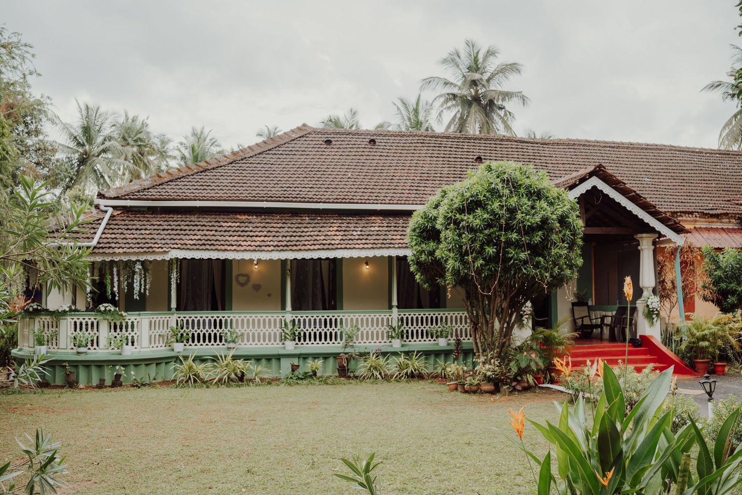 Single-story heritage villa with a tiled roof and white-railed veranda, surrounded by tall palm trees and tropical gardens.