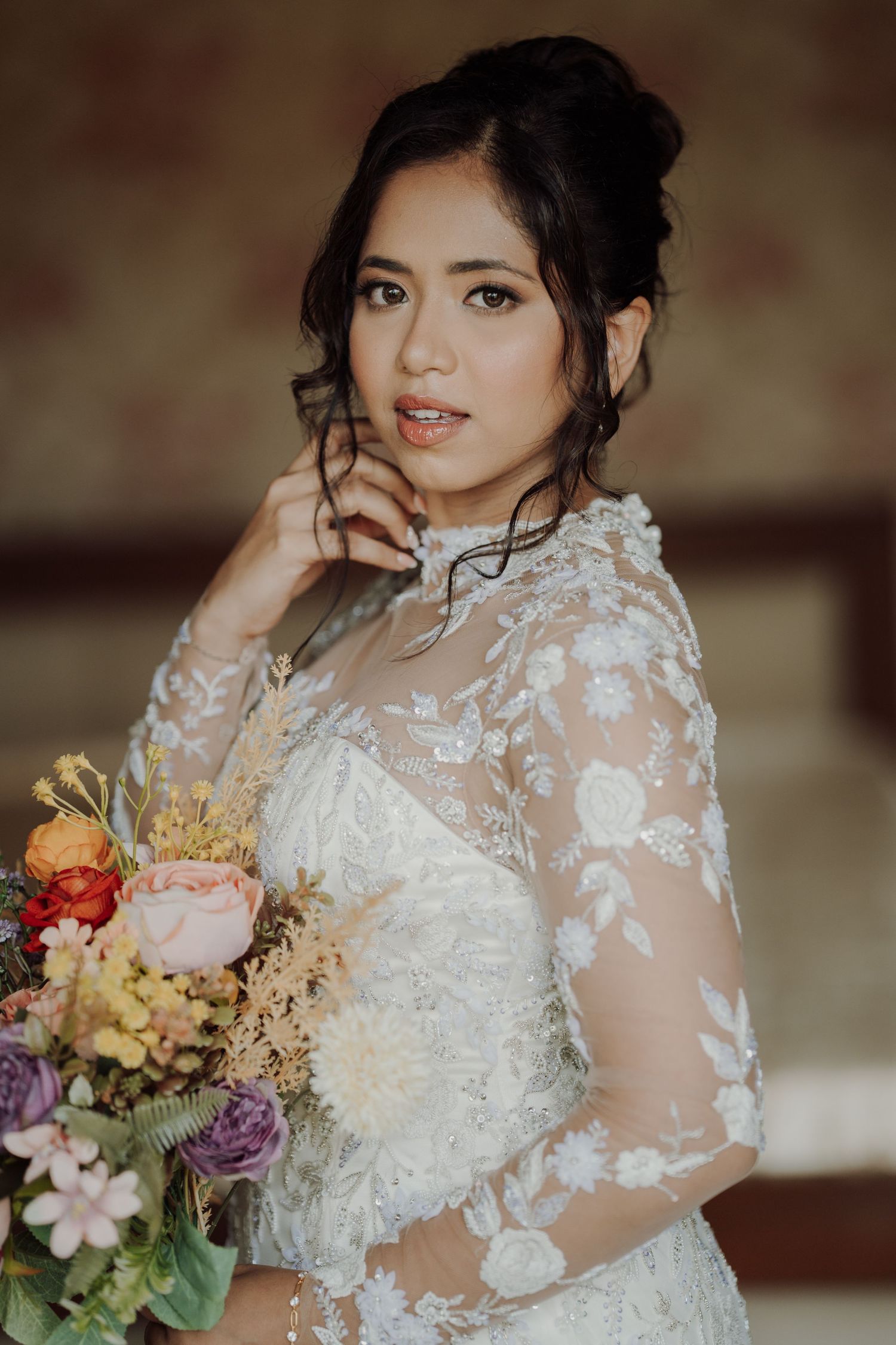 Bride in an elegant lace gown with floral embroidery and an updo holds a colorful bouquet of roses and wildflowers while posing indoors.