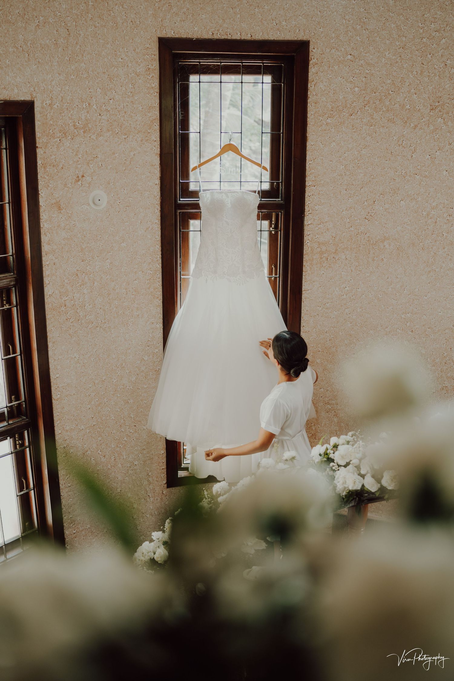 A white wedding dress hangs in front of a window while someone in a white robe sits nearby among white floral decorations.