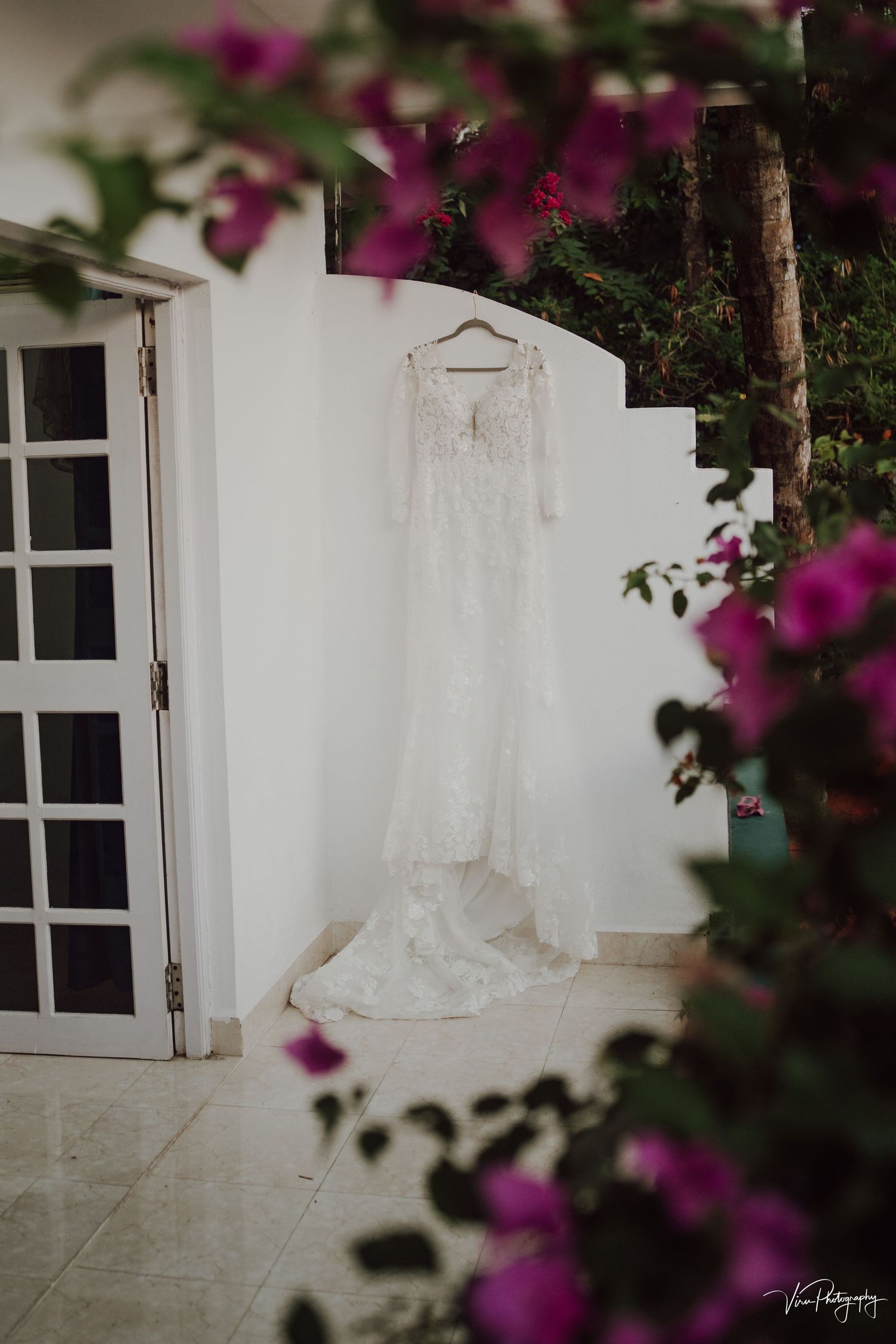 White lace wedding dress hangs elegantly against a white wall with blooming pink bougainvillea flowers framing the scene.