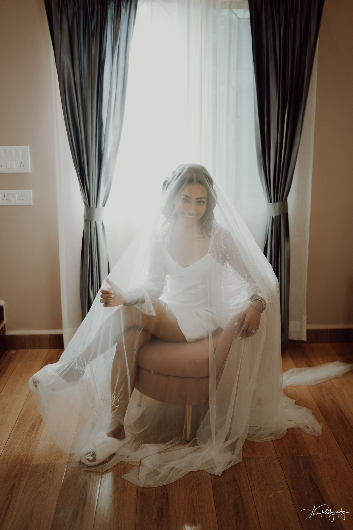 A bride in white robe sits elegantly on a beige chair by window with dark curtains, flowing veil creating ethereal atmosphere.