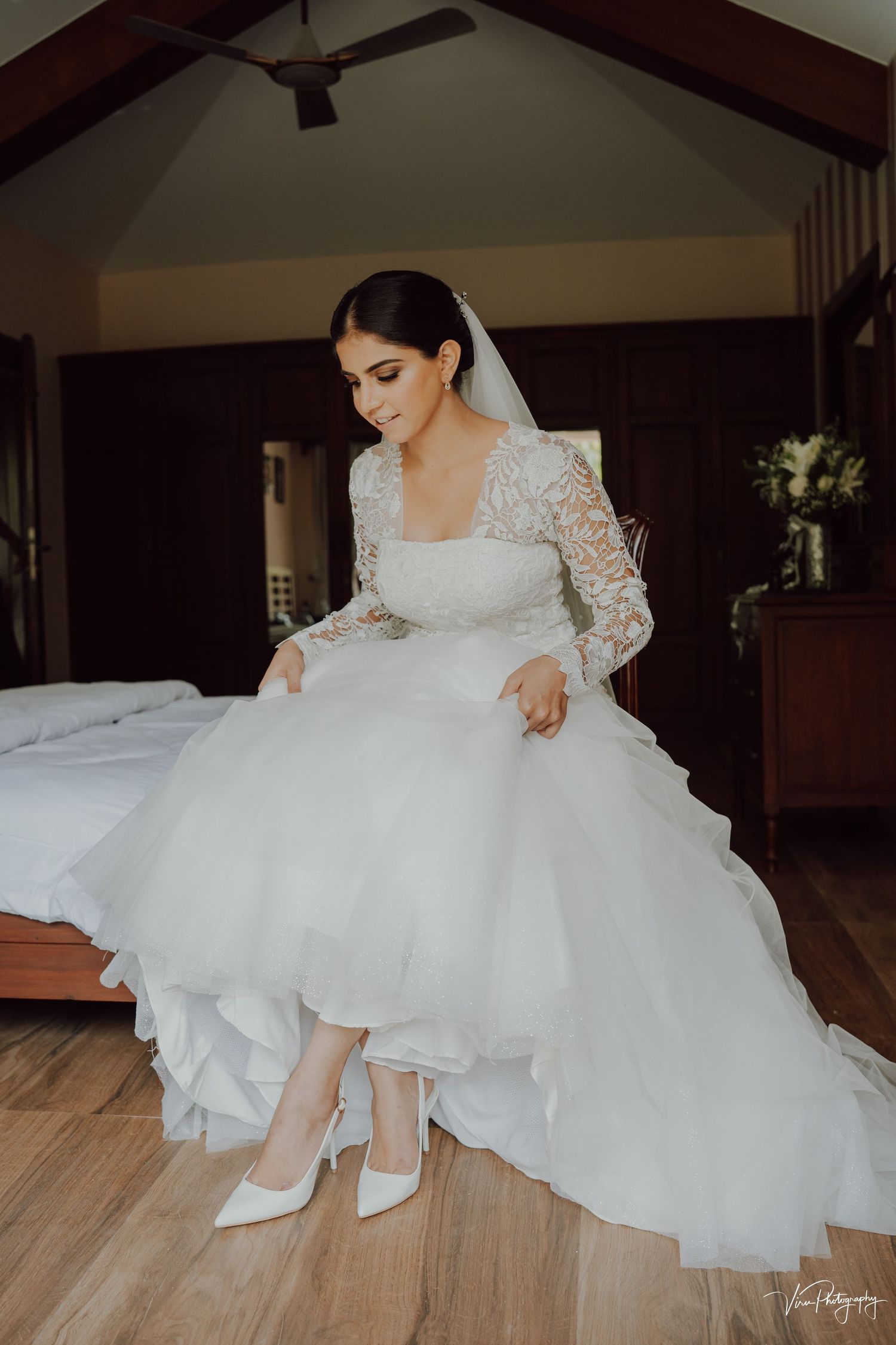 A bride in an elegant long-sleeved lace wedding gown prepares white heels while seated in a rustic wooden room.