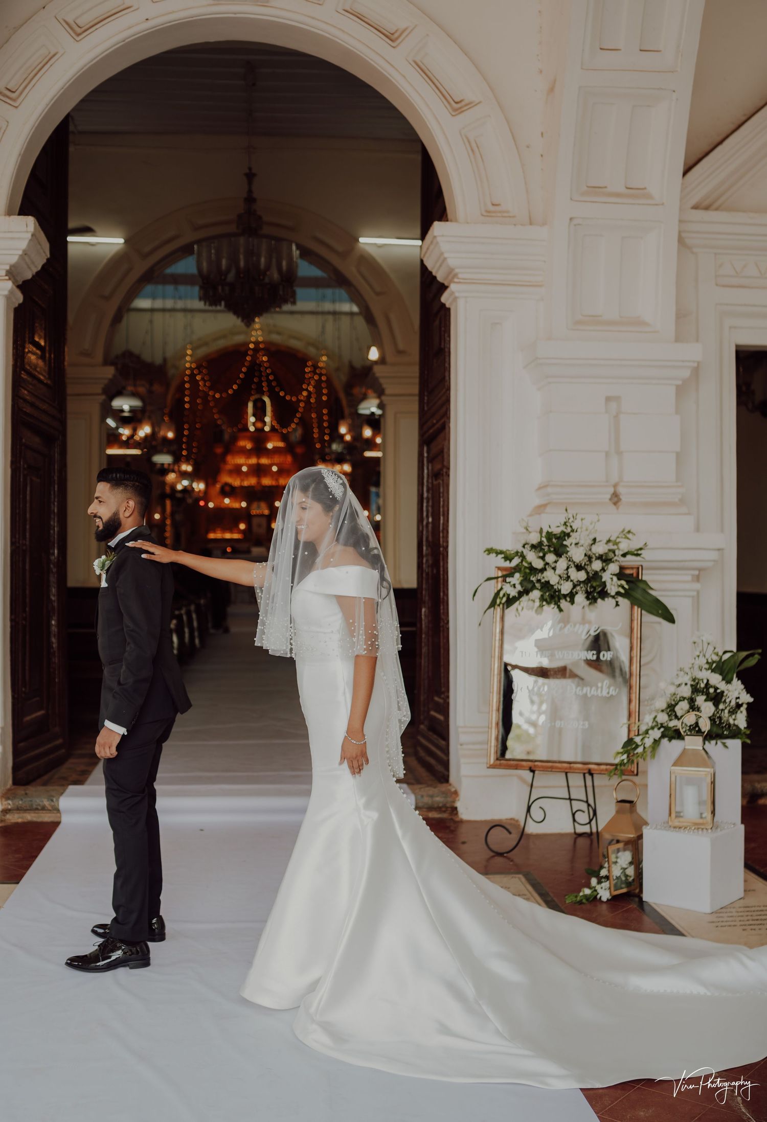 A wedding couple shares a moment under an elegant arched entrance decorated with white floral arrangements.