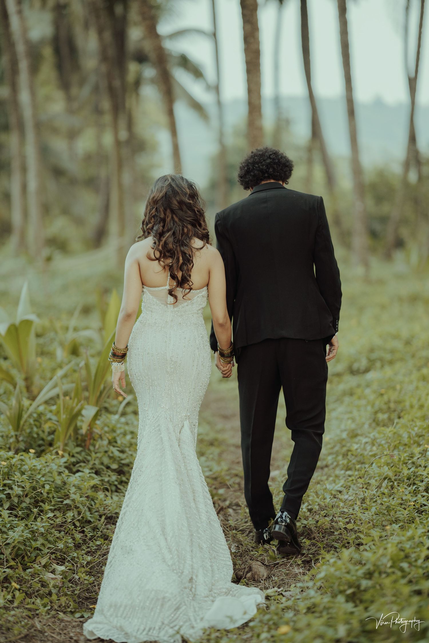Newlywed couple walking together on lush tropical path with palm trees and greenery in romantic wedding photo.