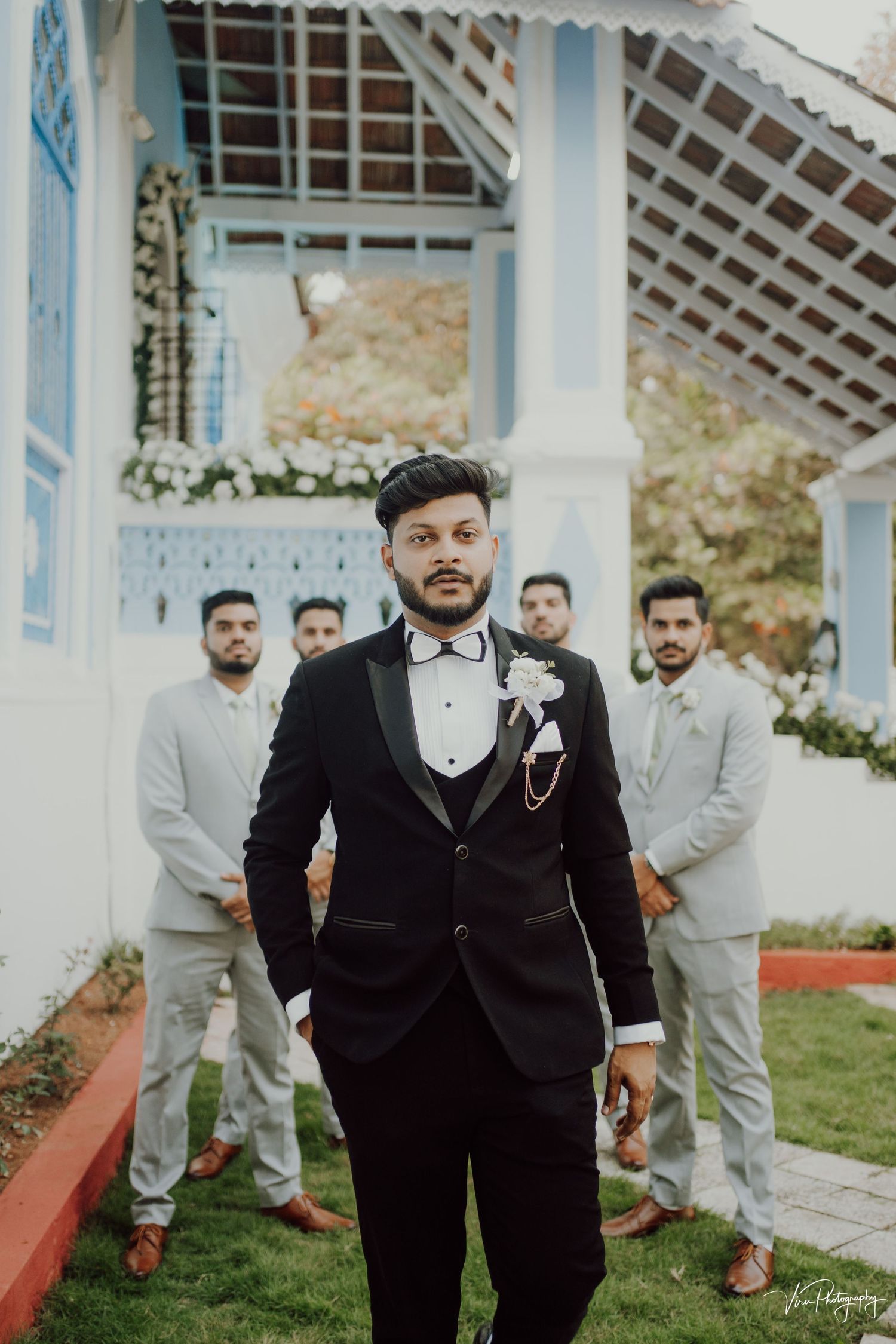 A wedding party poses at a white gazebo with a groom in black tuxedo and groomsmen wearing light gray suits.