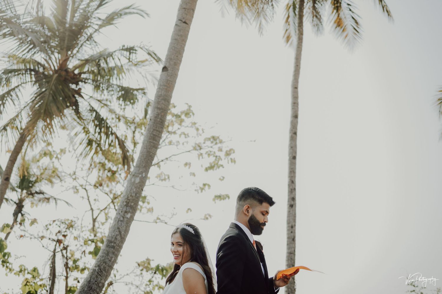 A couple stands beneath tall palm trees during a tropical destination wedding ceremony.