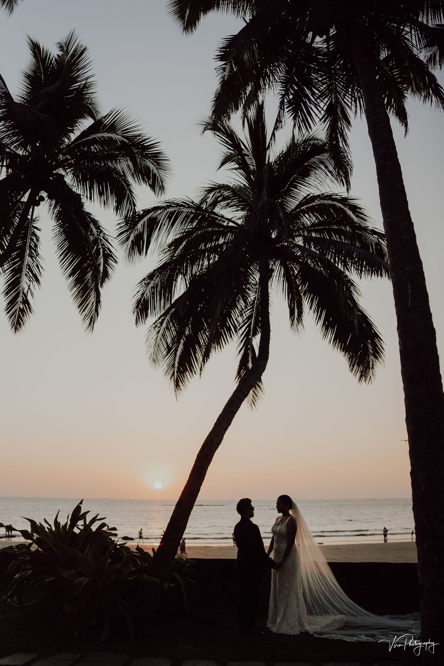 Silhouettes of palm trees frame a couple on a beach at sunset with gentle waves in the background.