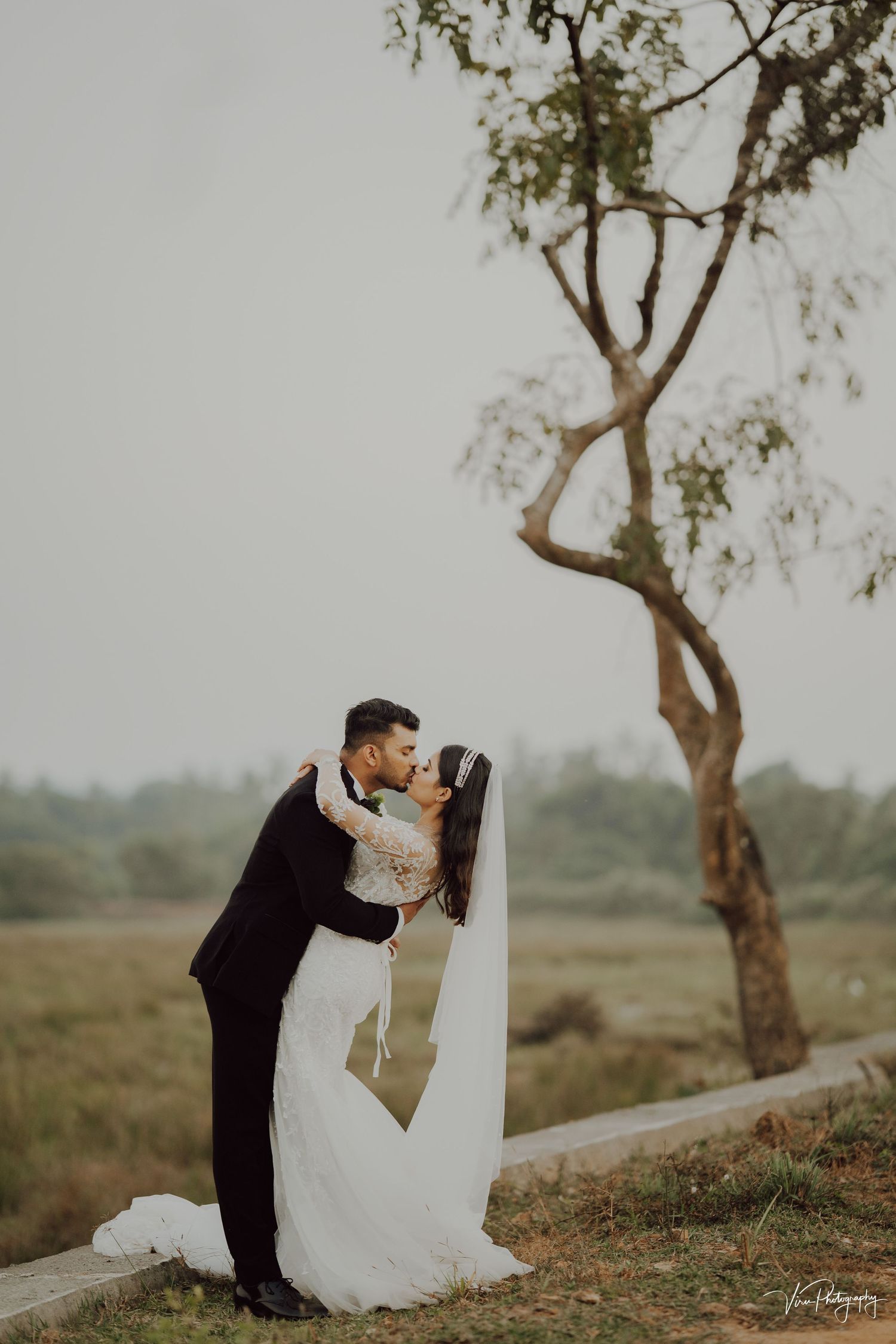 A couple shares a romantic kiss in the countryside under a twisted tree on their wedding day in warm, muted tones.