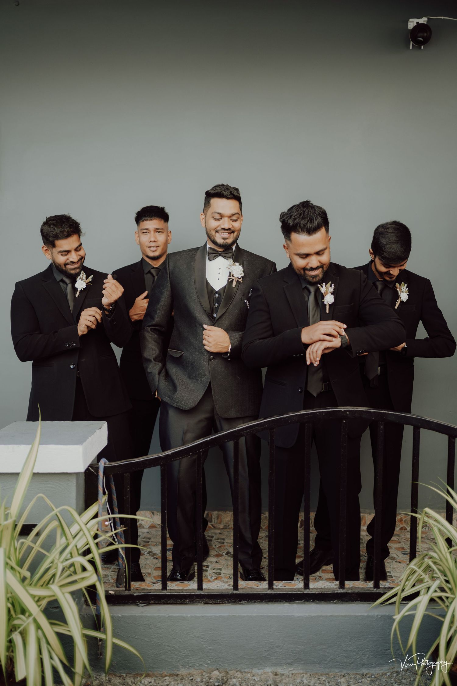 A group of groomsmen in black suits and bowties laugh together while standing on a porch with plants.