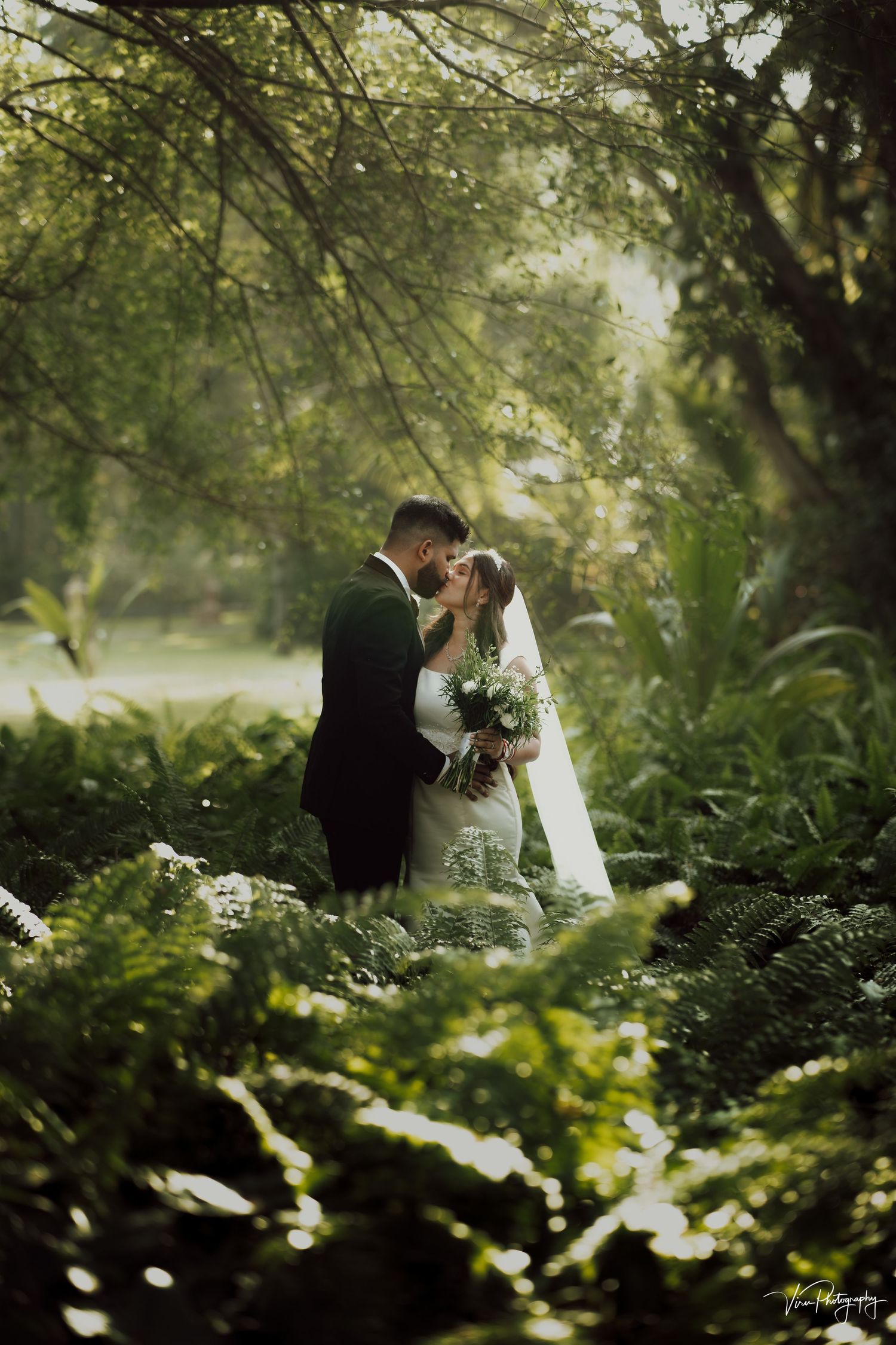 A romantic wedding portrait in a lush garden setting with sunlight filtering through green foliage.