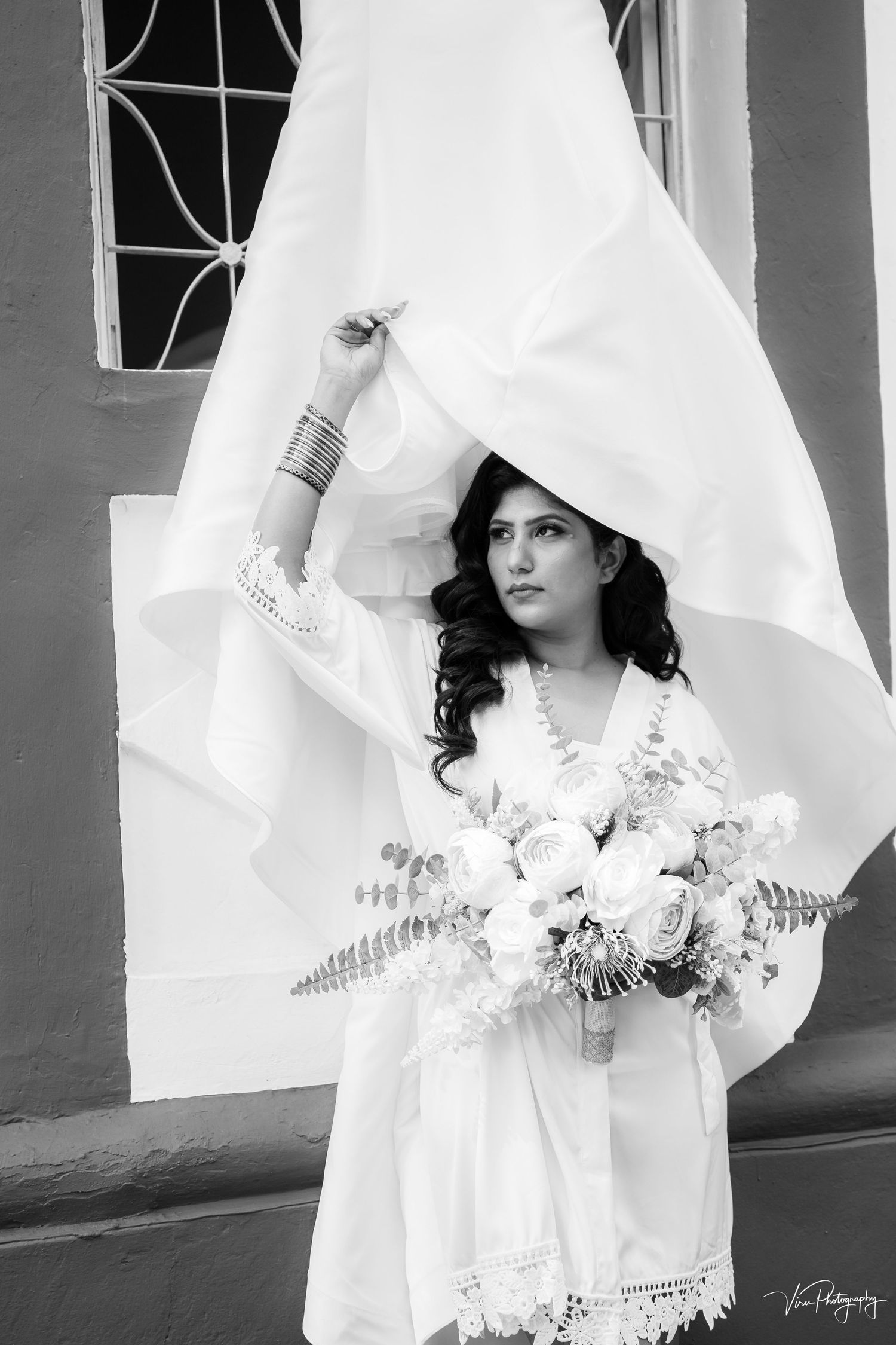 A bride in a flowing white dress holding a bouquet poses against a white backdrop in a black and white portrait.