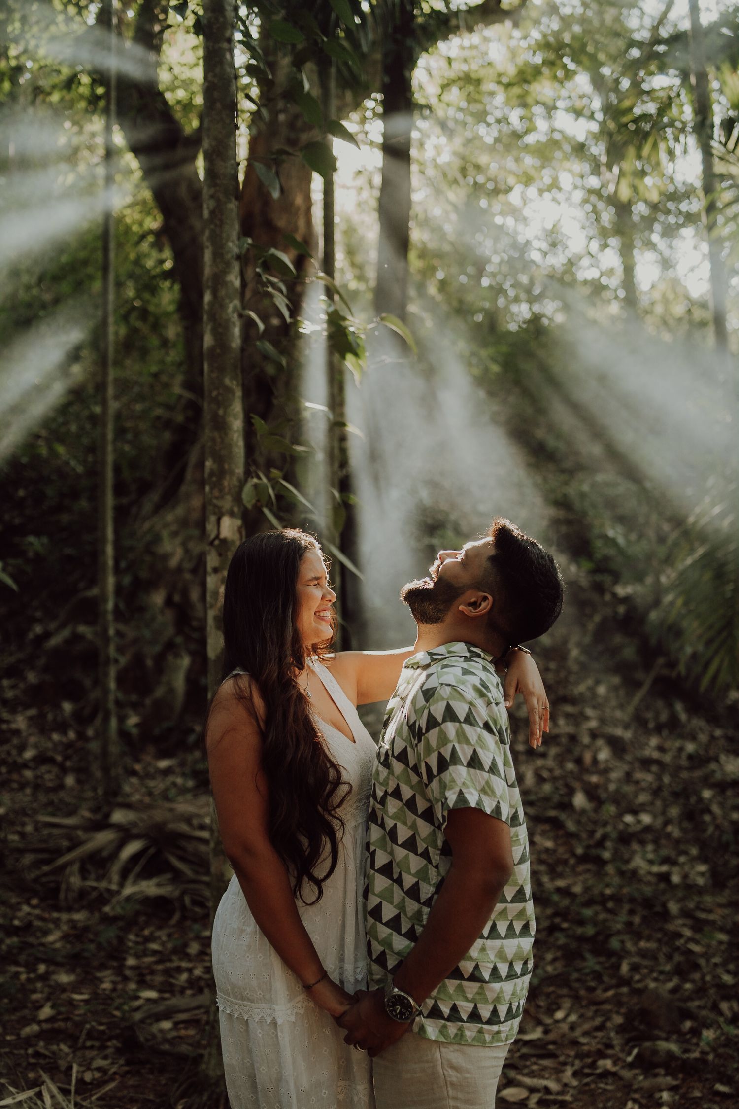 Couple hold hands and look up at sunbeams streaming through forest trees in ethereal woodland setting.