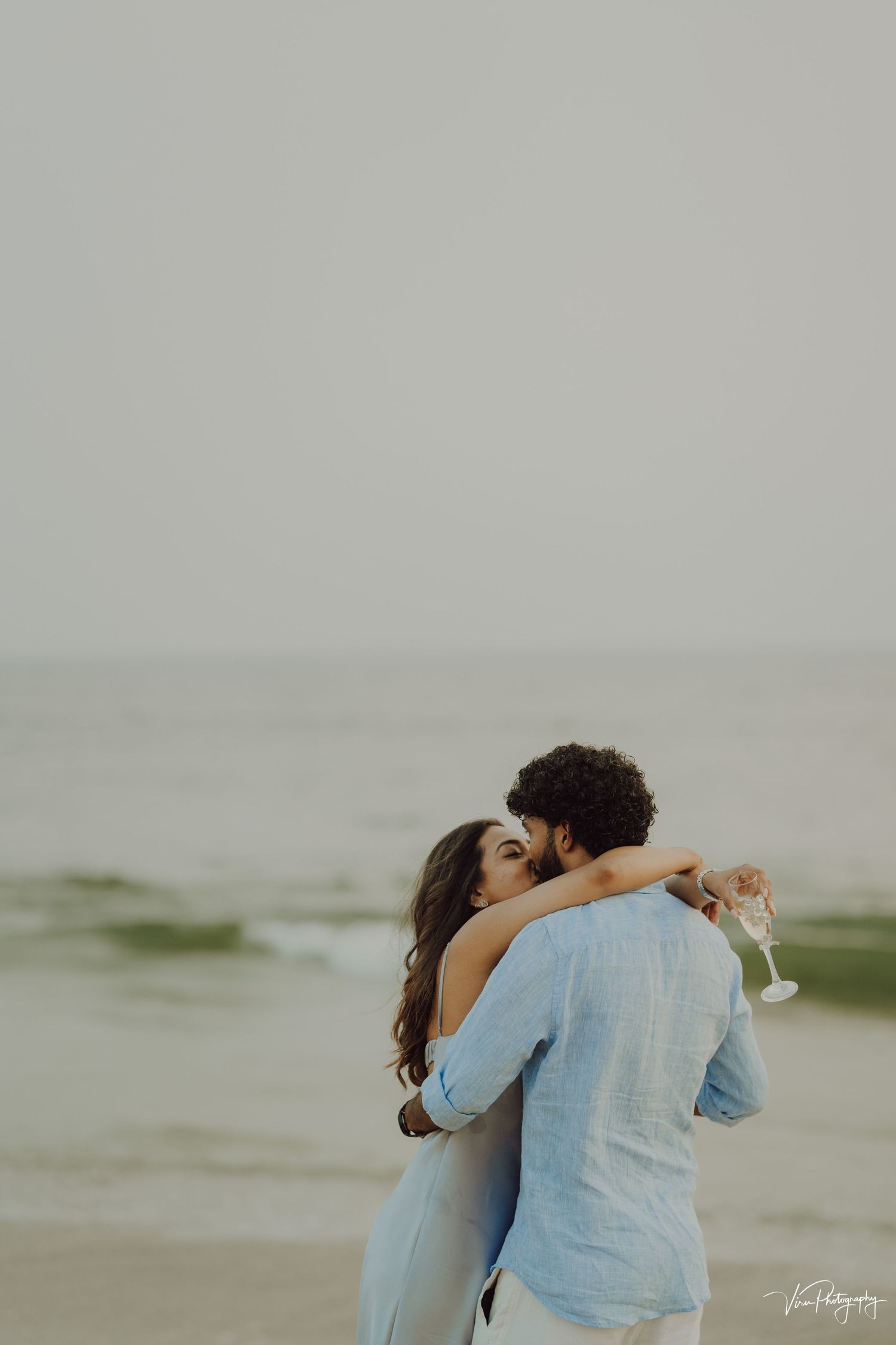 A romantic couple embraces and kisses on a misty beach with soft waves in the background.