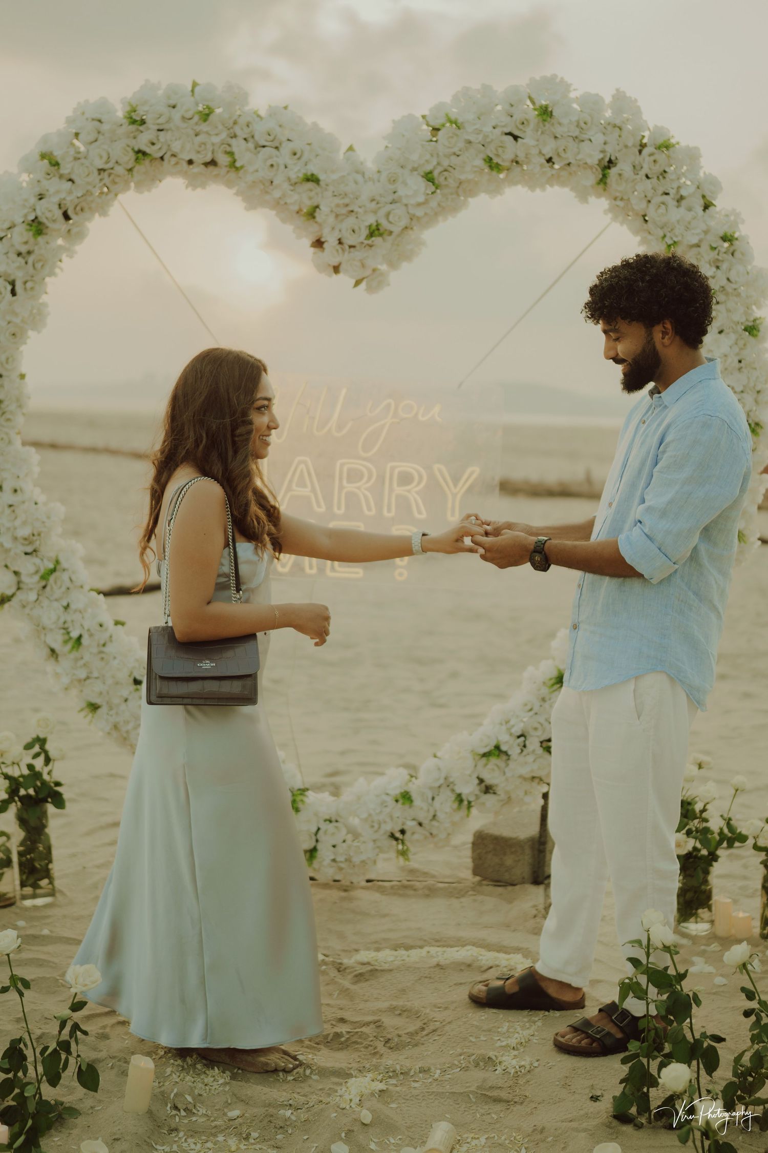 A romantic proposal scene under a floral arch by the beach at sunset with soft lighting and natural coastal backdrop.