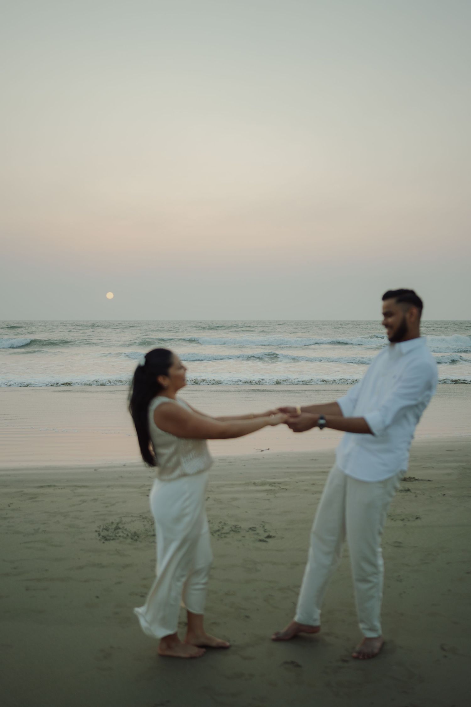 Couple holding hands on a beach at sunset with ocean waves and a full moon in the background.