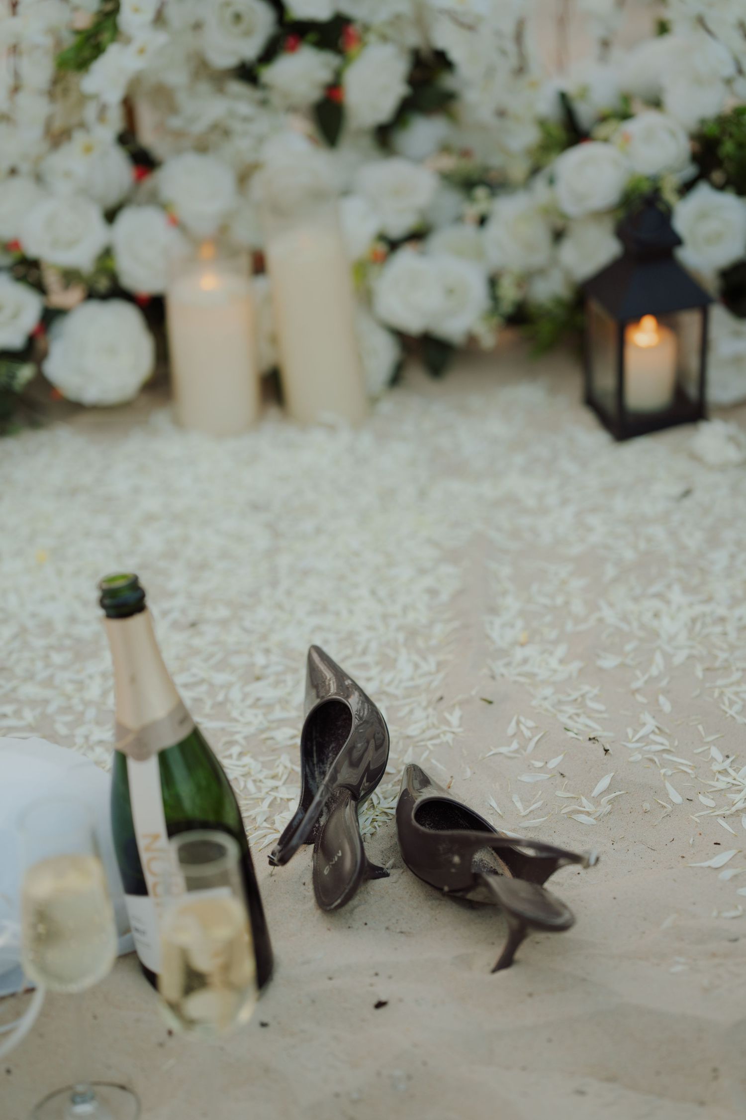 Champagne bottle and glasses beside discarded heels on sandy beach with white floral arrangements and candles in background.