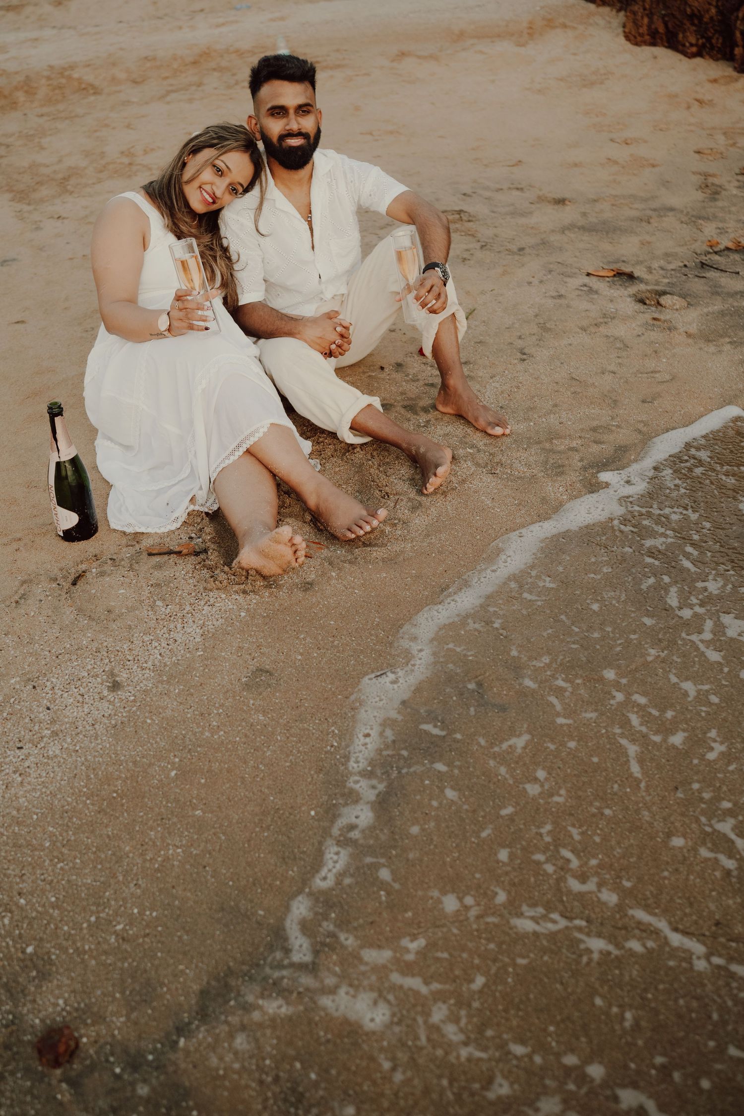 A couple in white outfits sits together on a sandy beach as gentle waves wash over their bare feet.