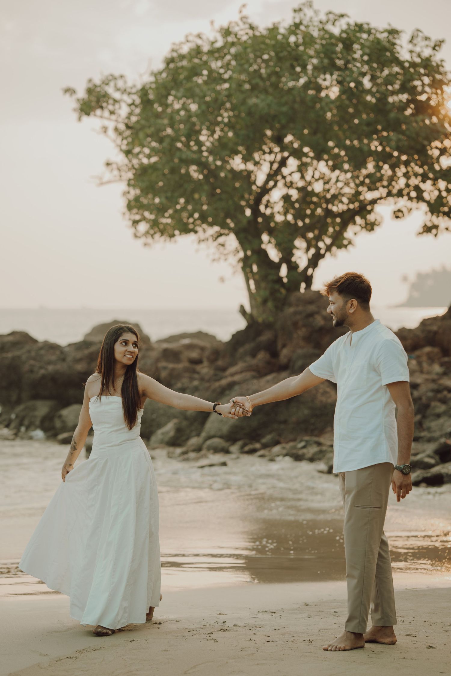 A couple holds hands on a beach at sunset with a large tree silhouetted against the sky in the background.