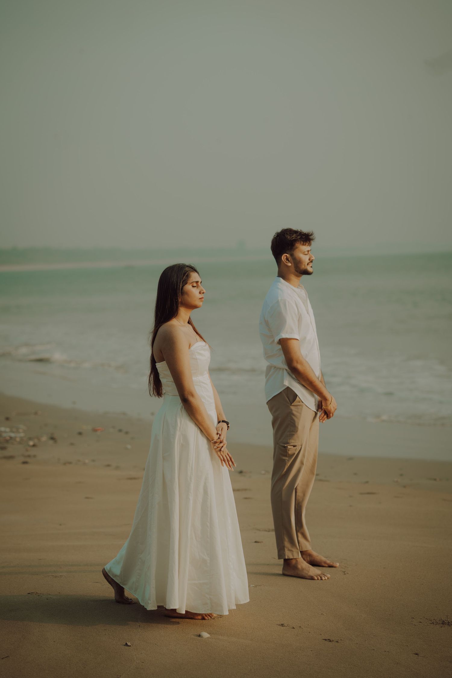 A couple stands back-to-back on a misty beach, wearing white and beige outfits during a romantic photoshoot.