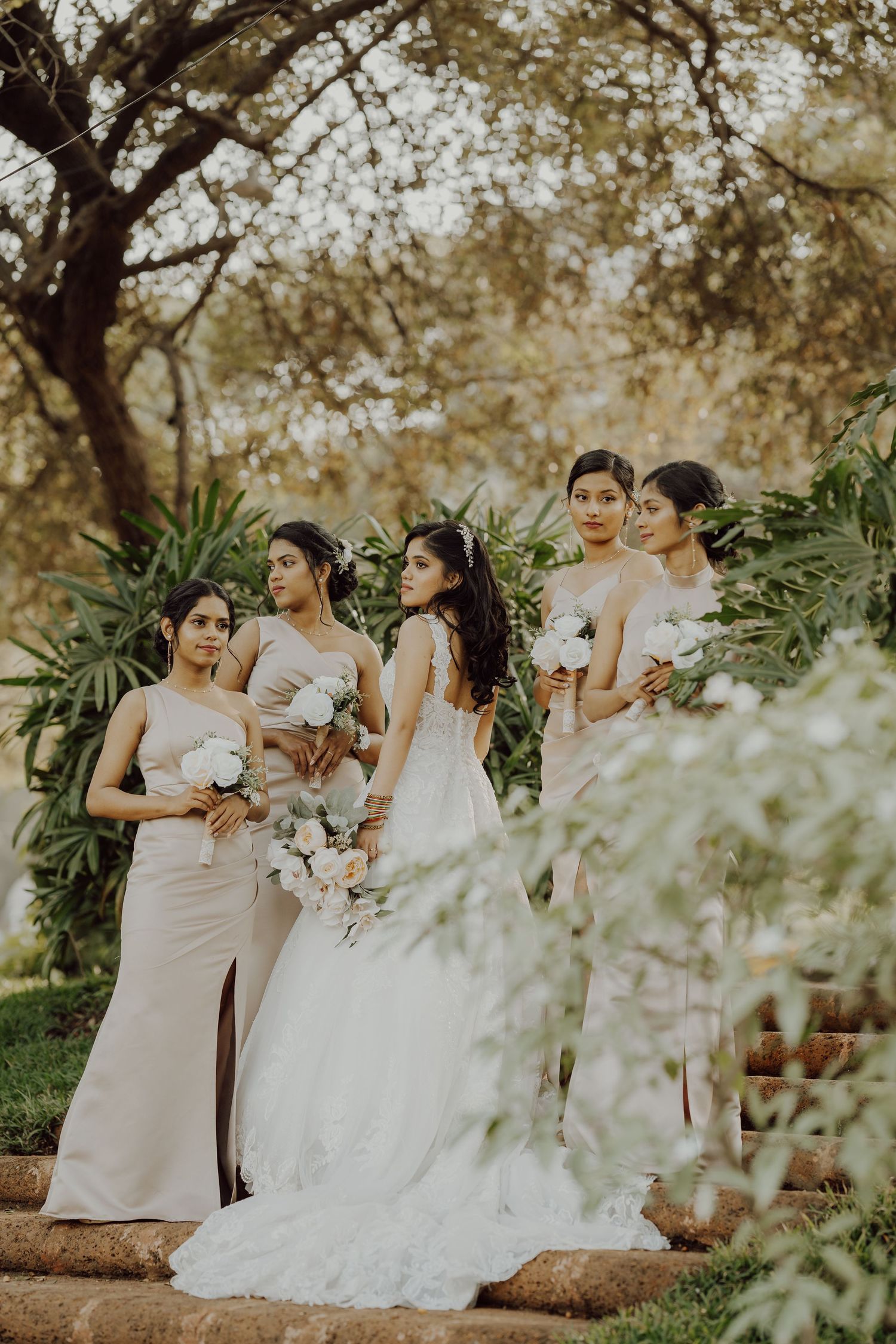 Elegant bridal party poses in white gowns amidst lush garden foliage.