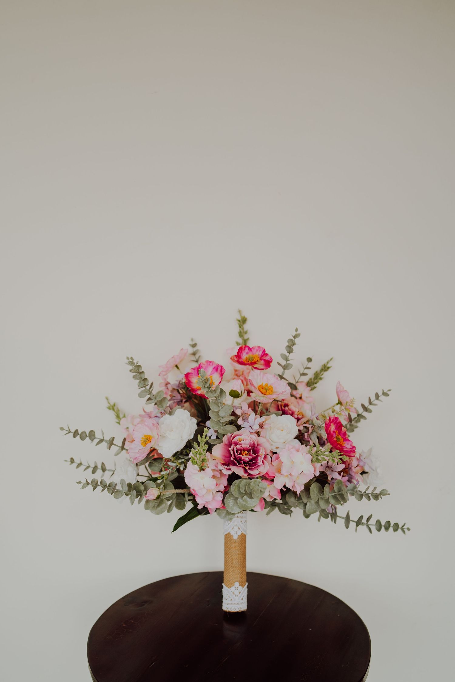 A romantic floral bouquet with pink roses, white flowers and eucalyptus sits on a dark round table against a neutral wall.