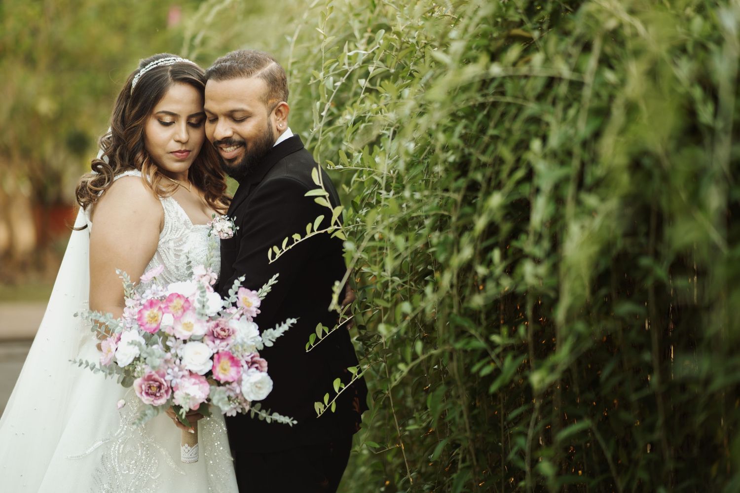 A romantic wedding moment with pink and white bouquet as they embrace near lush greenery in a garden setting.
