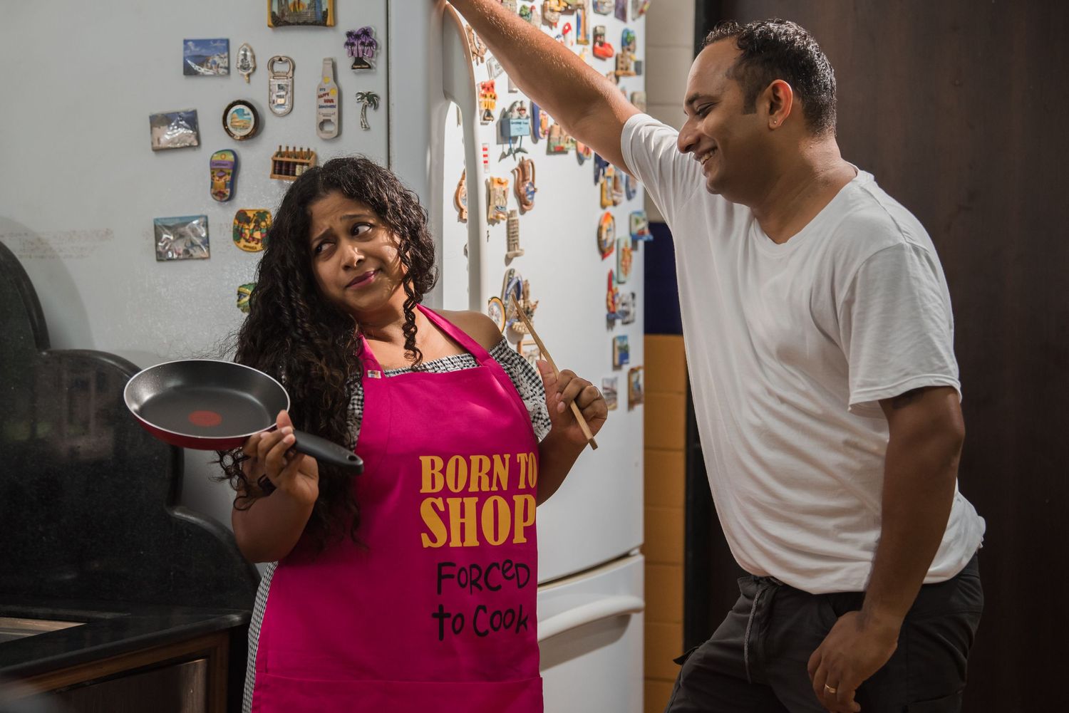 Two people cook together in a kitchen, with one wearing a bright pink apron that says 'Born 2 Shop'.