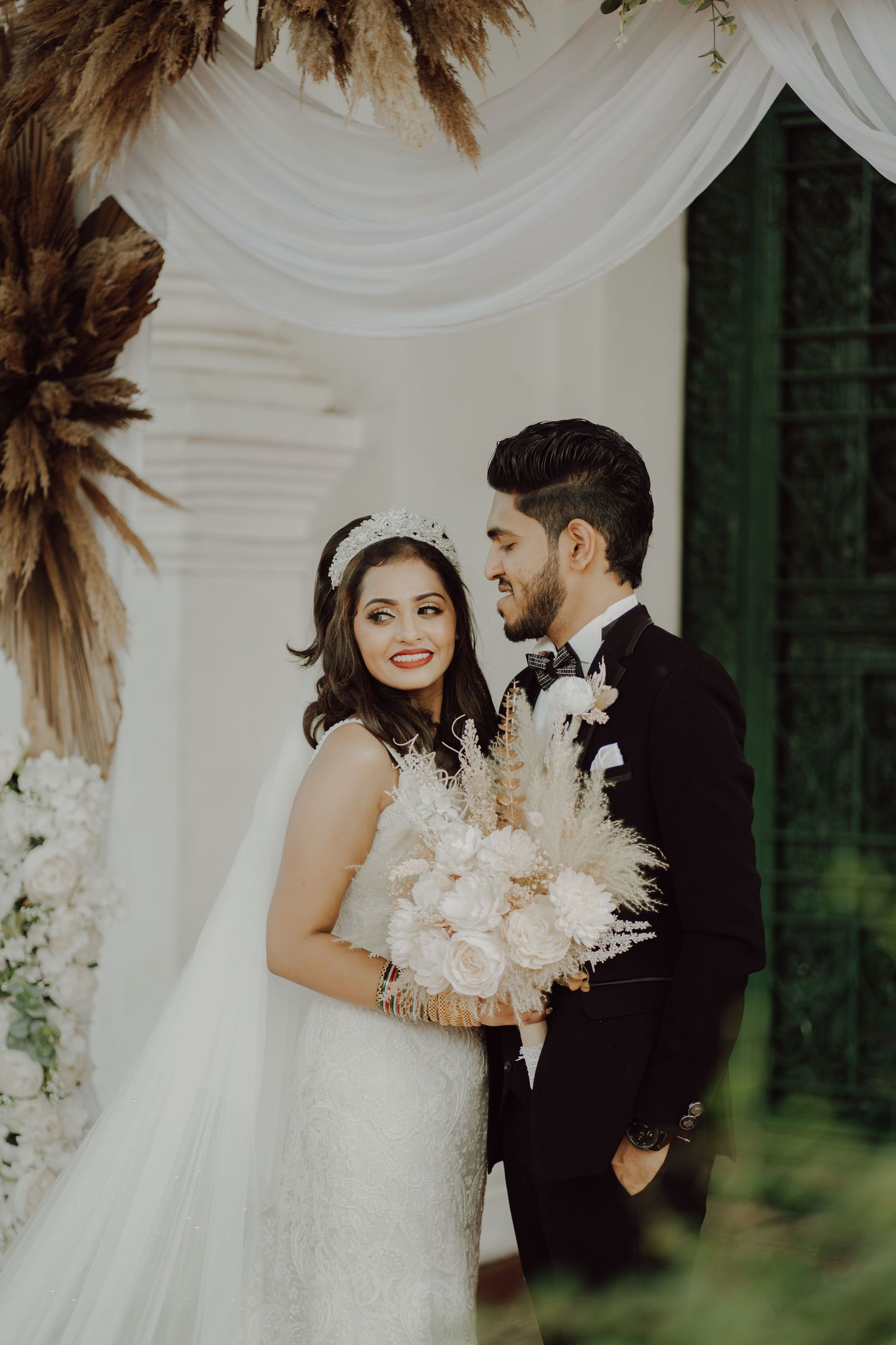 Newlywed couple in elegant attire pose under white drapery and dried pampas grass wedding arch decoration.