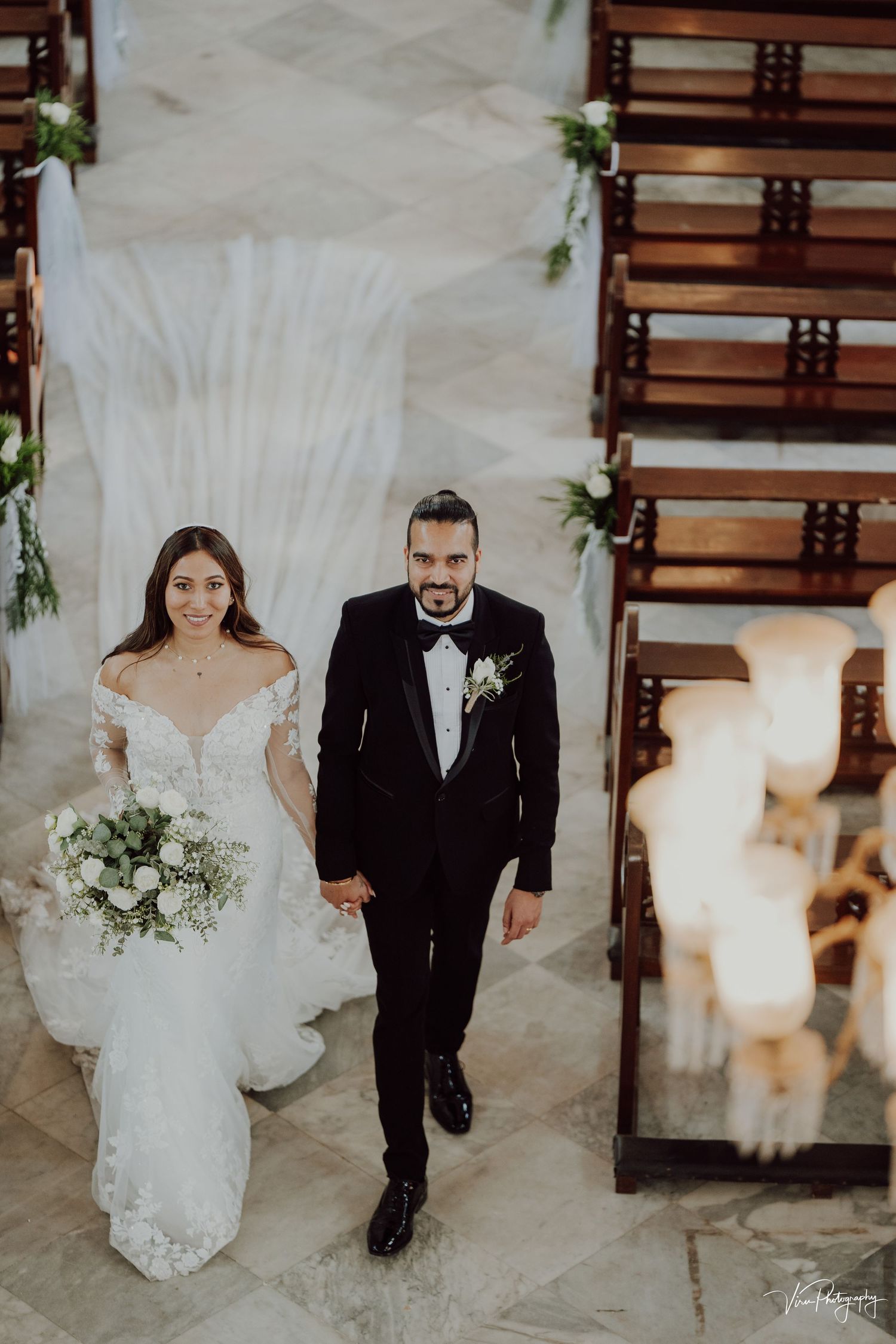 A bride in an off-shoulder lace gown and groom in a black tuxedo walk together at their elegant indoor wedding ceremony.