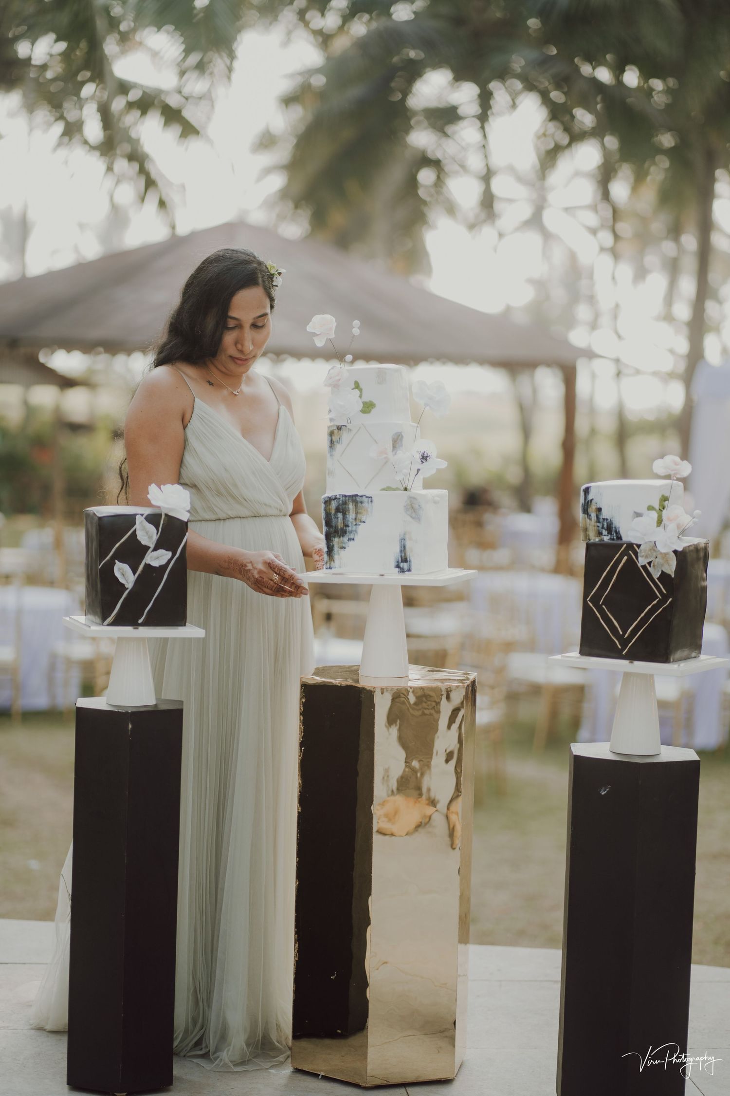 A dreamy outdoor wedding setup features elegant white floral arrangements displayed on black pedestals with metallic accents.