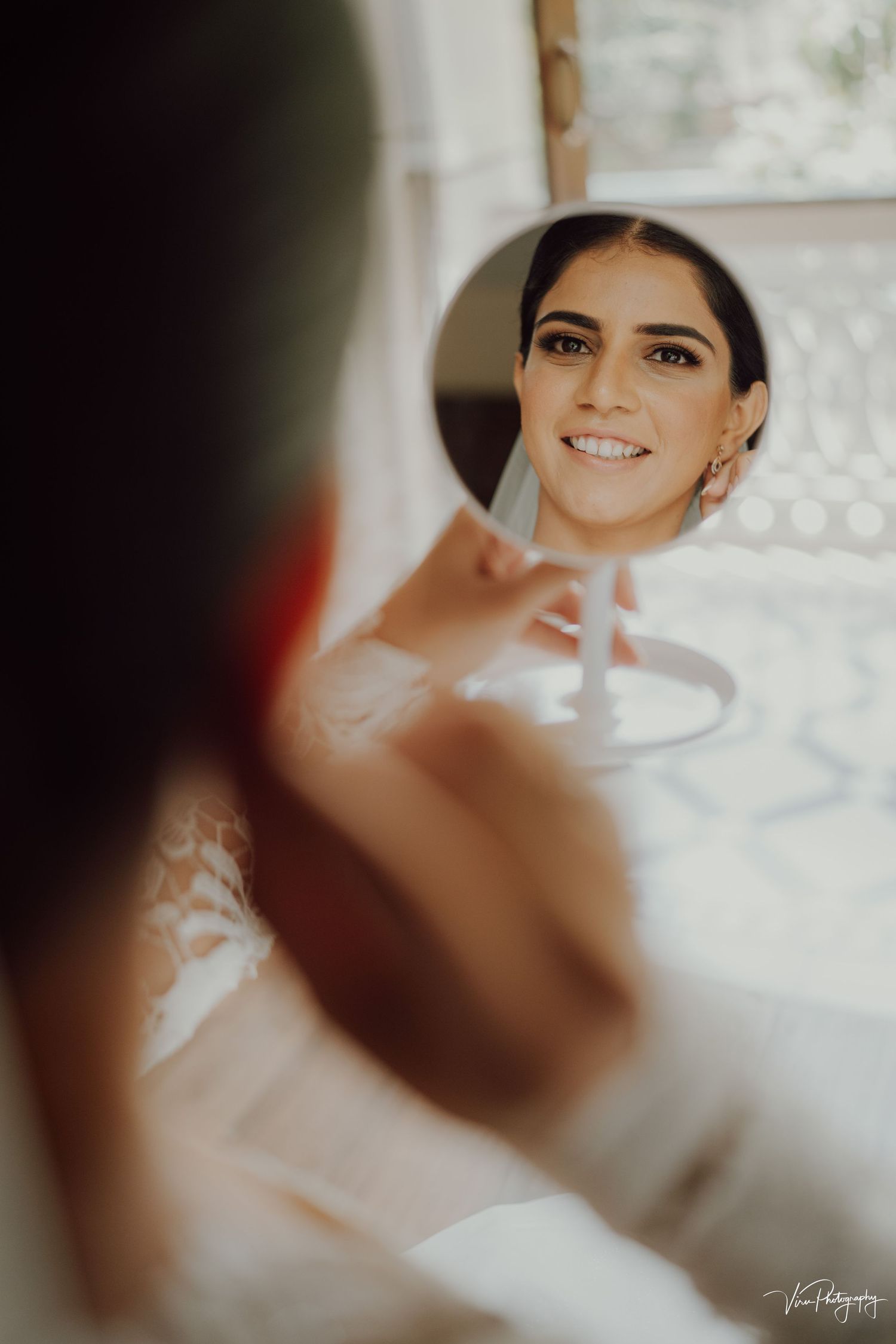 Reflection in mirror shows bride getting ready with a radiant smile while wearing a white wedding dress.