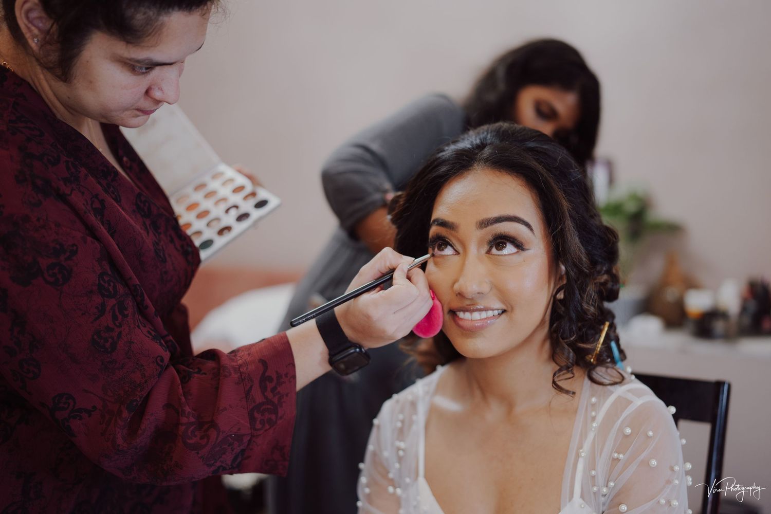 A makeup artist applies cosmetics on someone wearing a white lace dress during wedding preparations.
