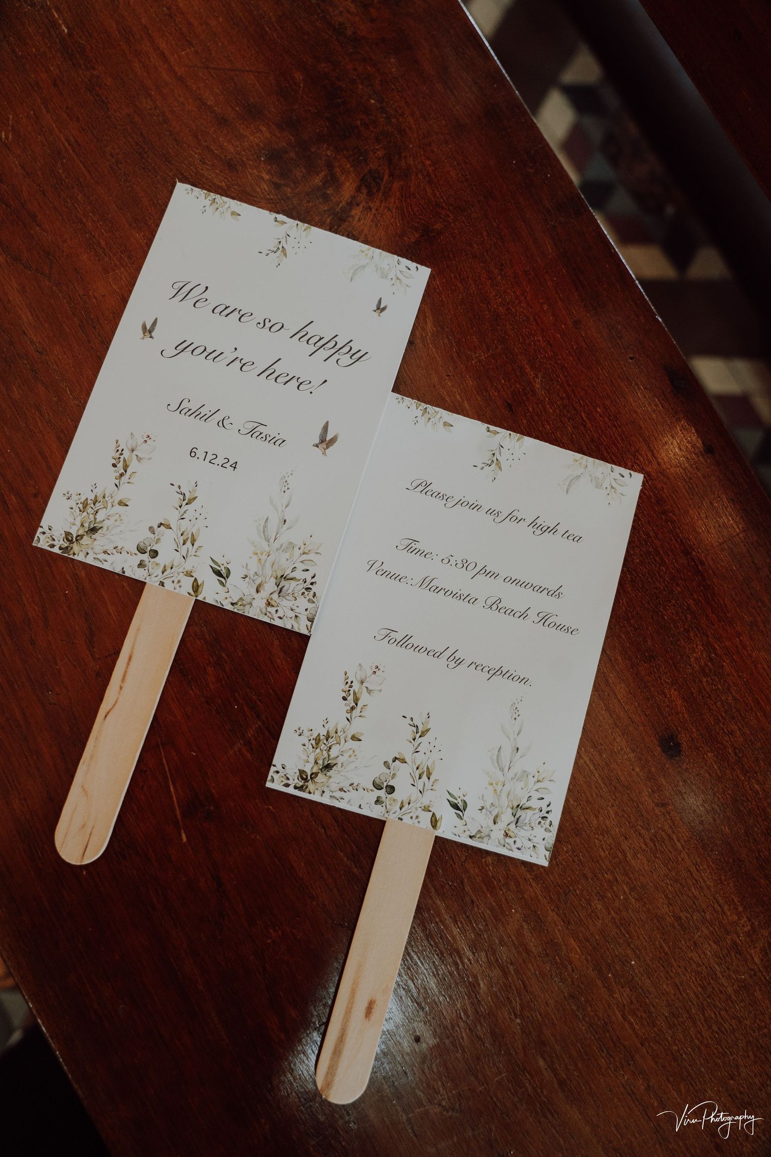 Wedding program fans with delicate floral designs printed on white paper with wooden handles on dark wooden surface.