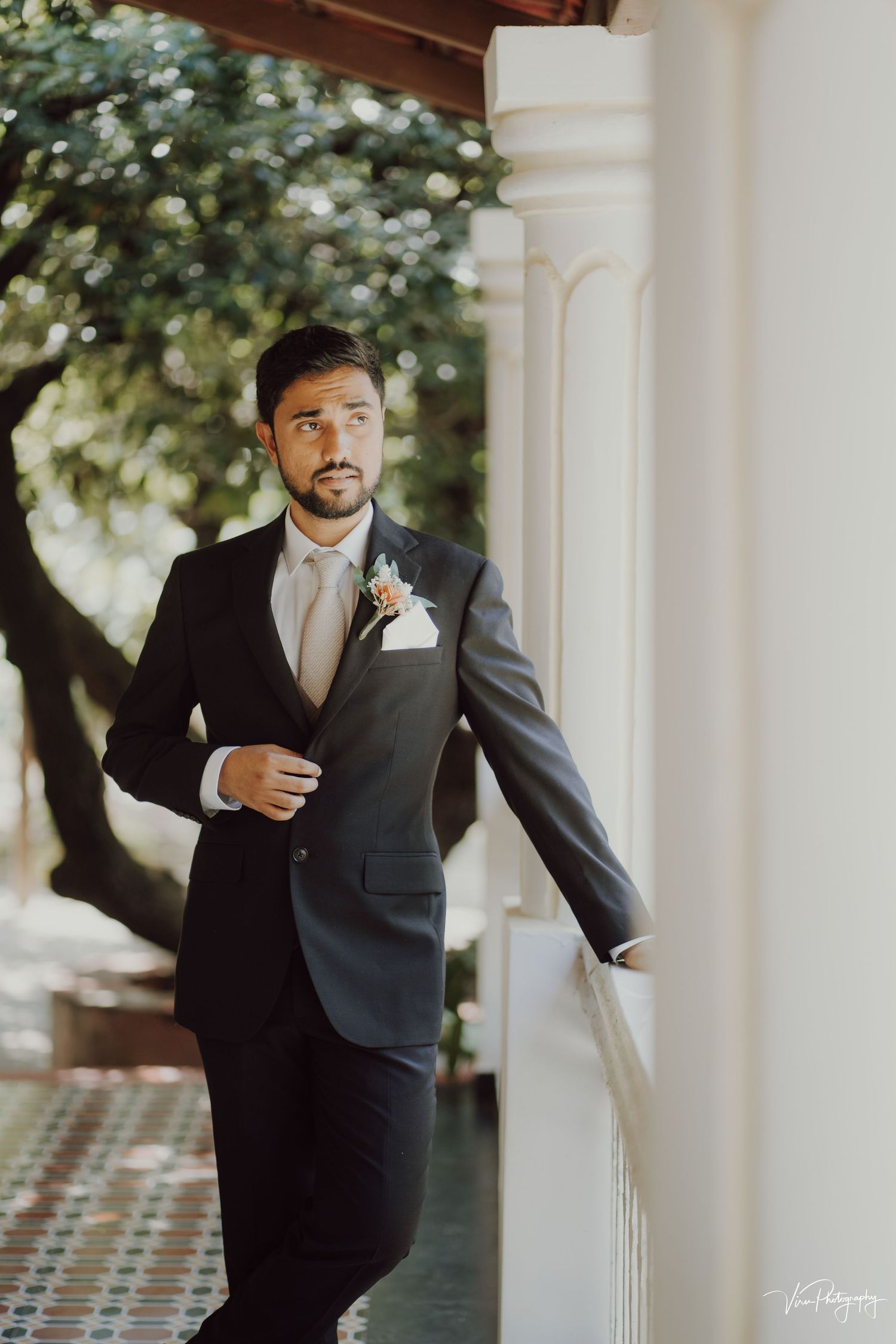 Groom in classic black suit and tie poses confidently against white column during outdoor wedding photoshoot.