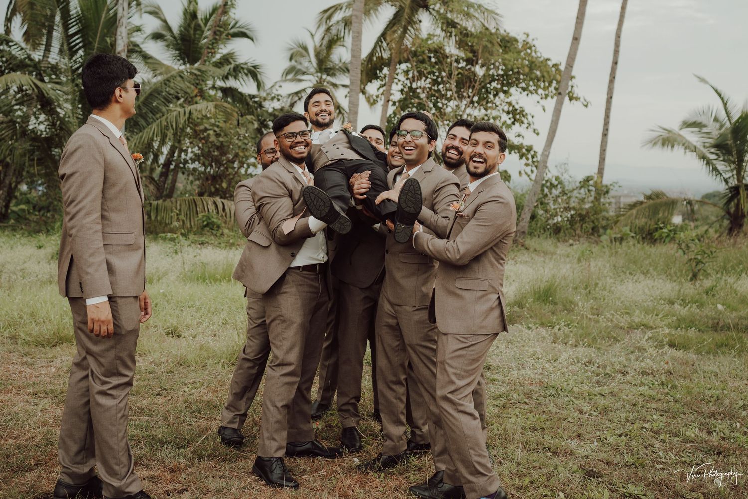 A candid moment shows a wedding party in tan suits playfully lifting and celebrating in an outdoor tropical setting.