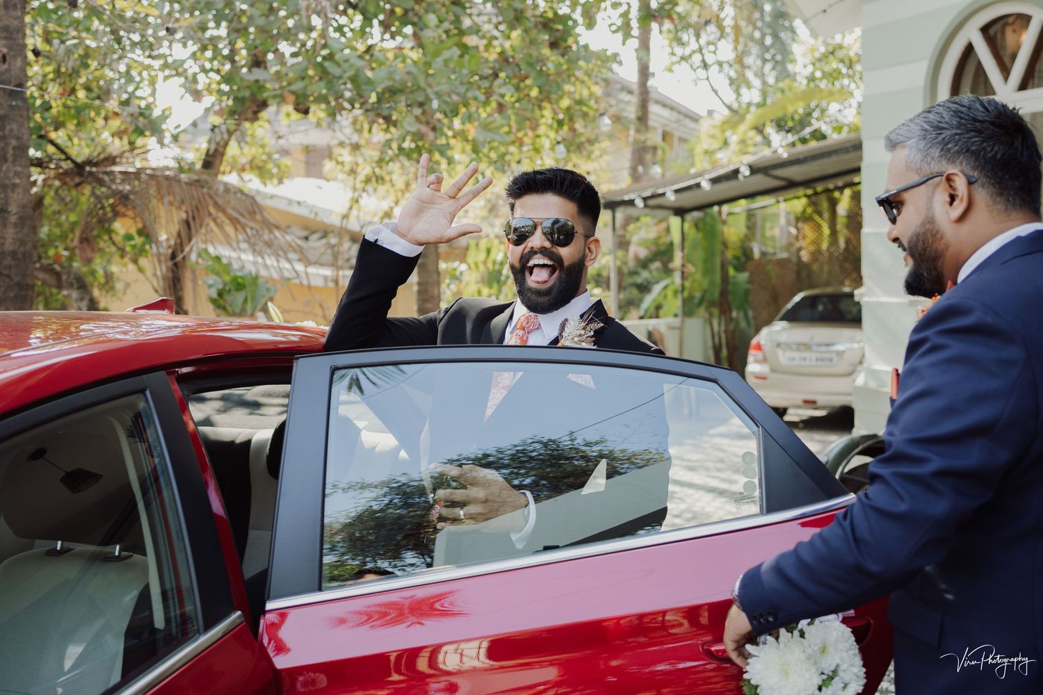 Two well-dressed men in business attire stand by a red car, with one waving from the open passenger door.