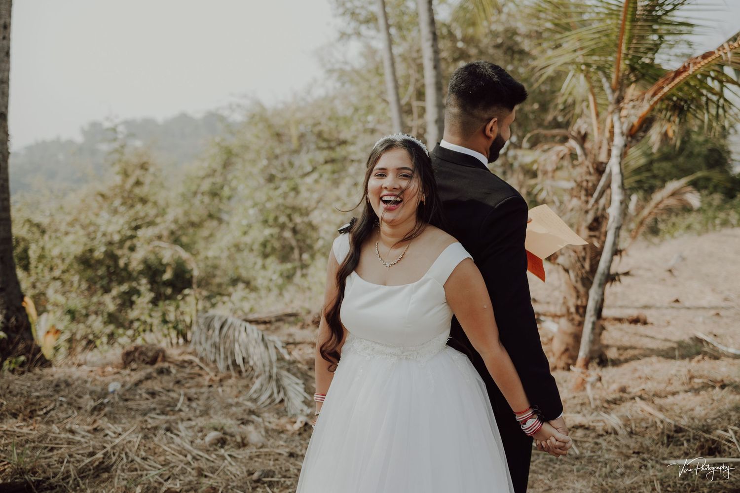 A couple in wedding attire share a joyful moment on a hilltop surrounded by tropical foliage.