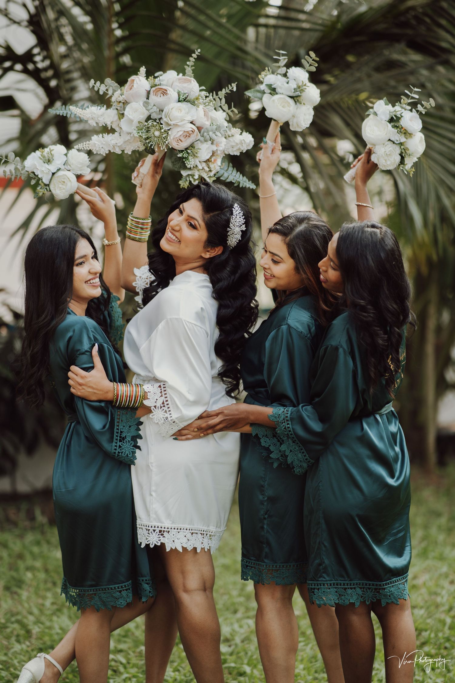 Bride in white robe celebrates with bridesmaids in teal silk robes holding white floral bouquets in a tropical garden setting.