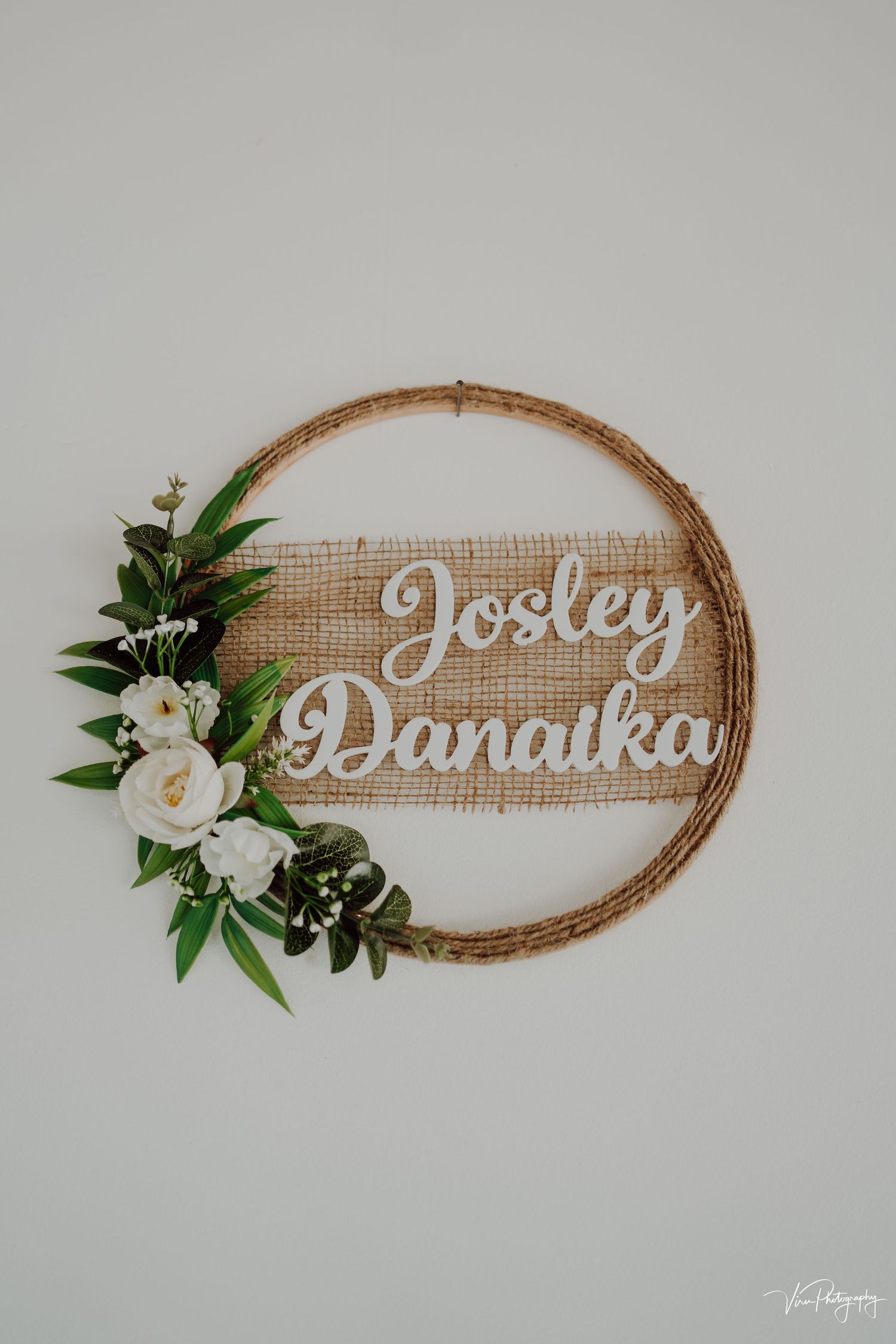 A rustic wooden hoop wreath with burlap fabric, white roses, greenery and cursive text reading 'Josley Danaika'.