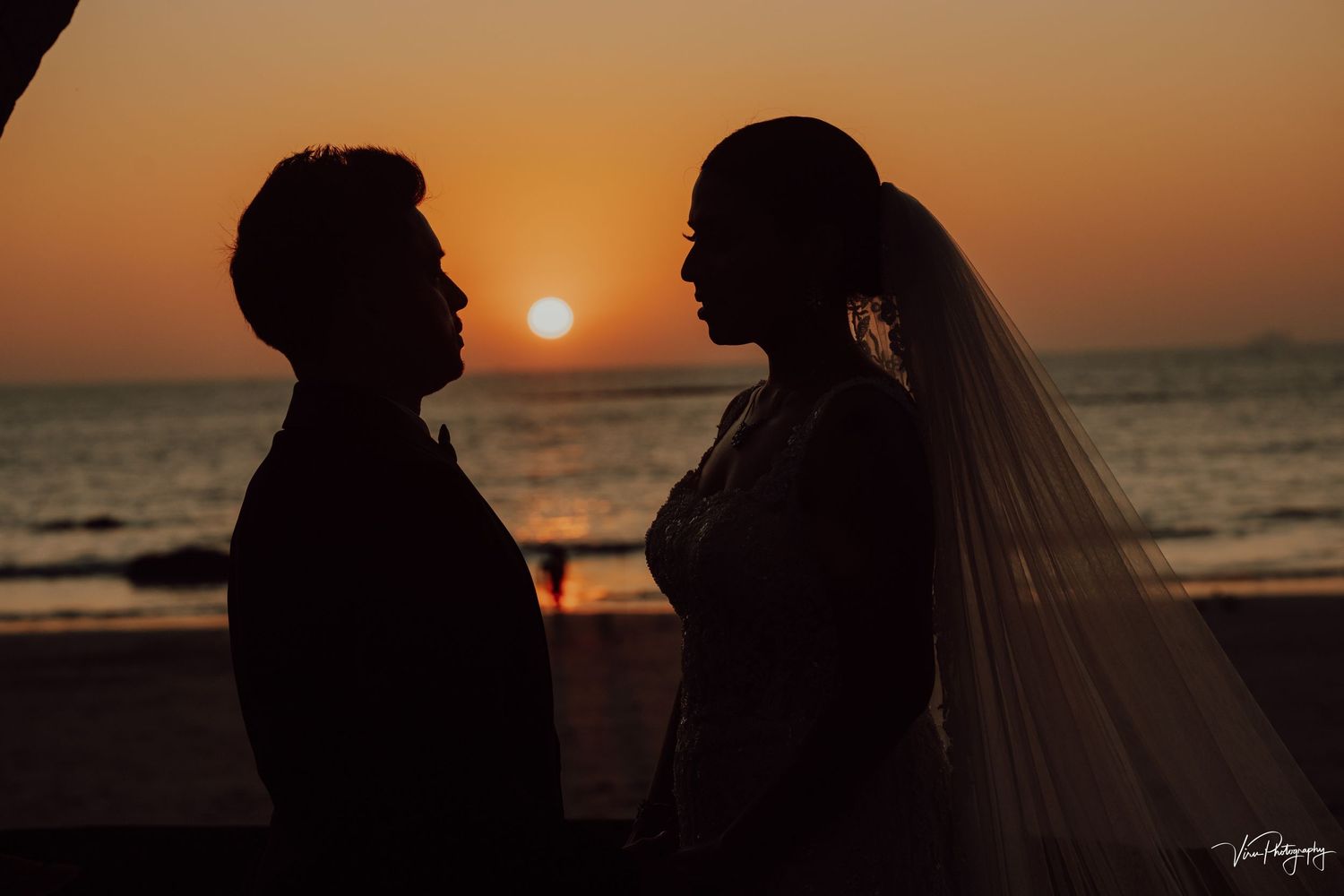 Silhouettes of two romantic figures stand together against an orange sunset over the ocean beach as a wedding veil flows in the breeze.