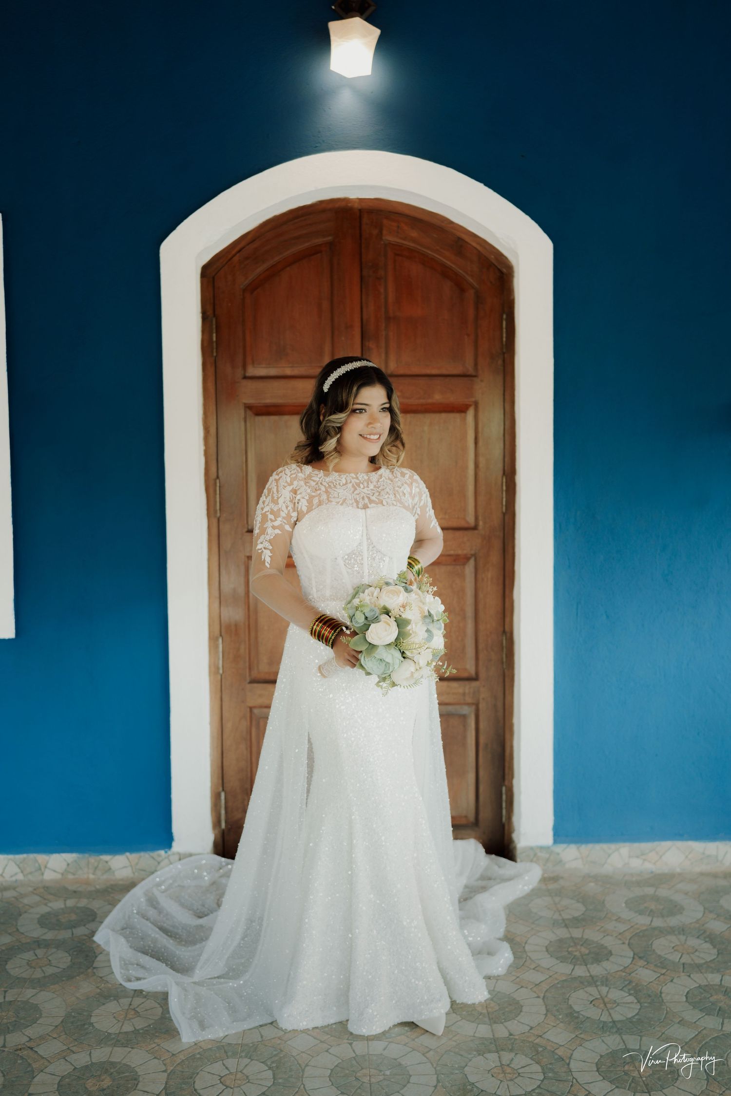 A bride in a white lace wedding dress with sleeves stands in front of a wooden door against a vibrant blue wall.