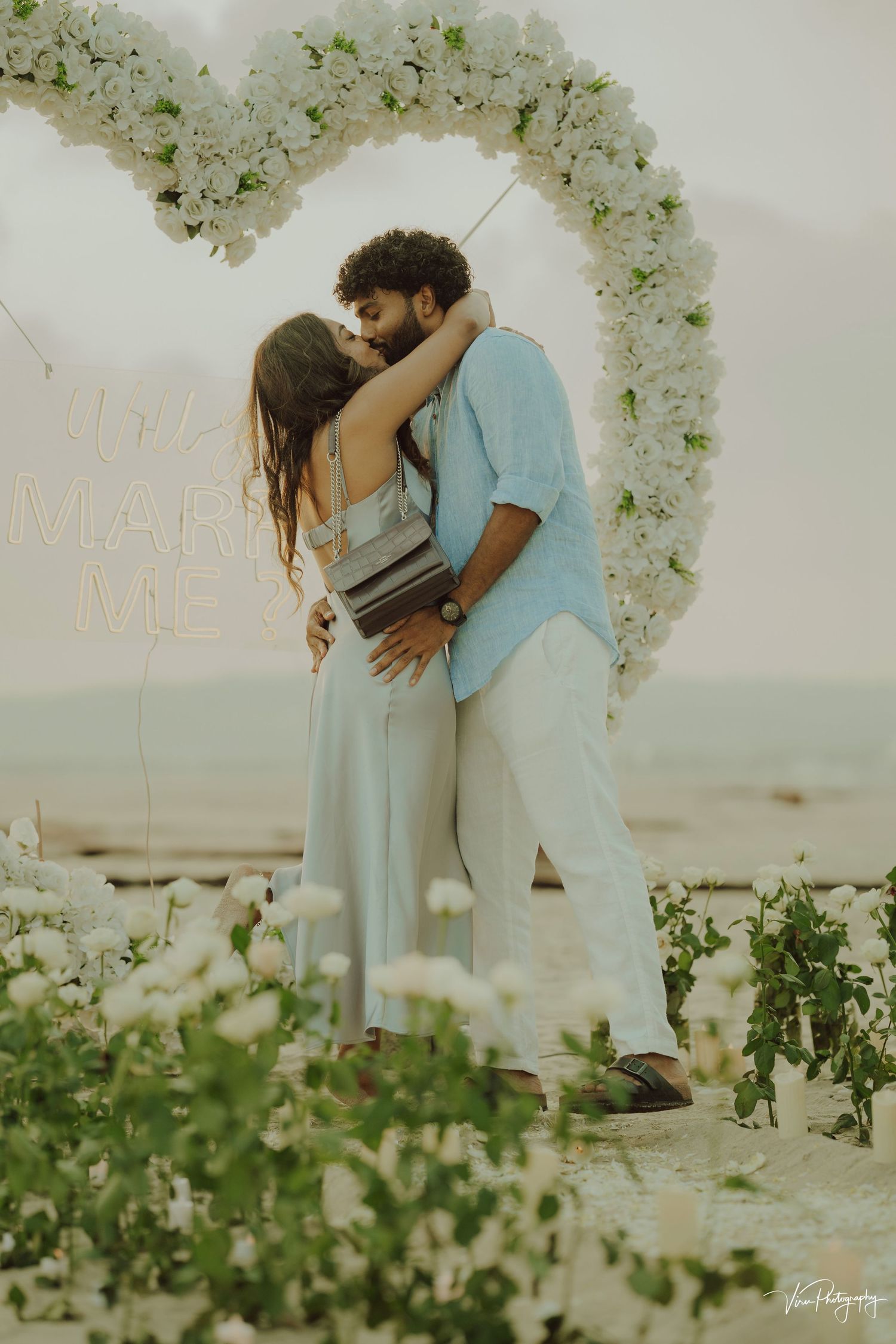 A romantic couple embraces under a white floral arch on a beach at golden hour during a marriage proposal.