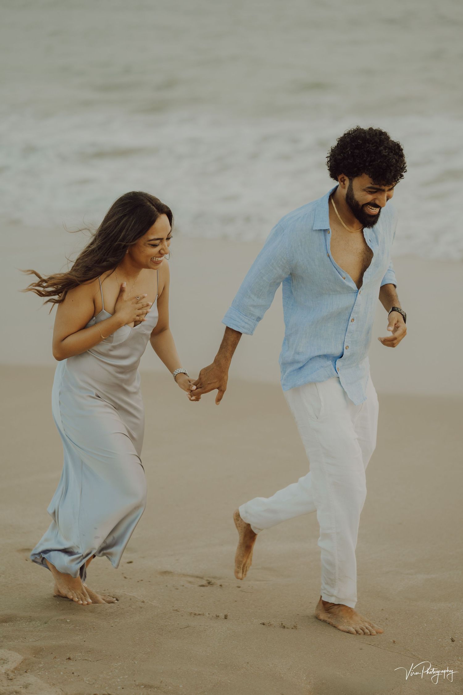 A couple holds hands while running together on a beach, wearing light flowy outfits in a romantic scene.