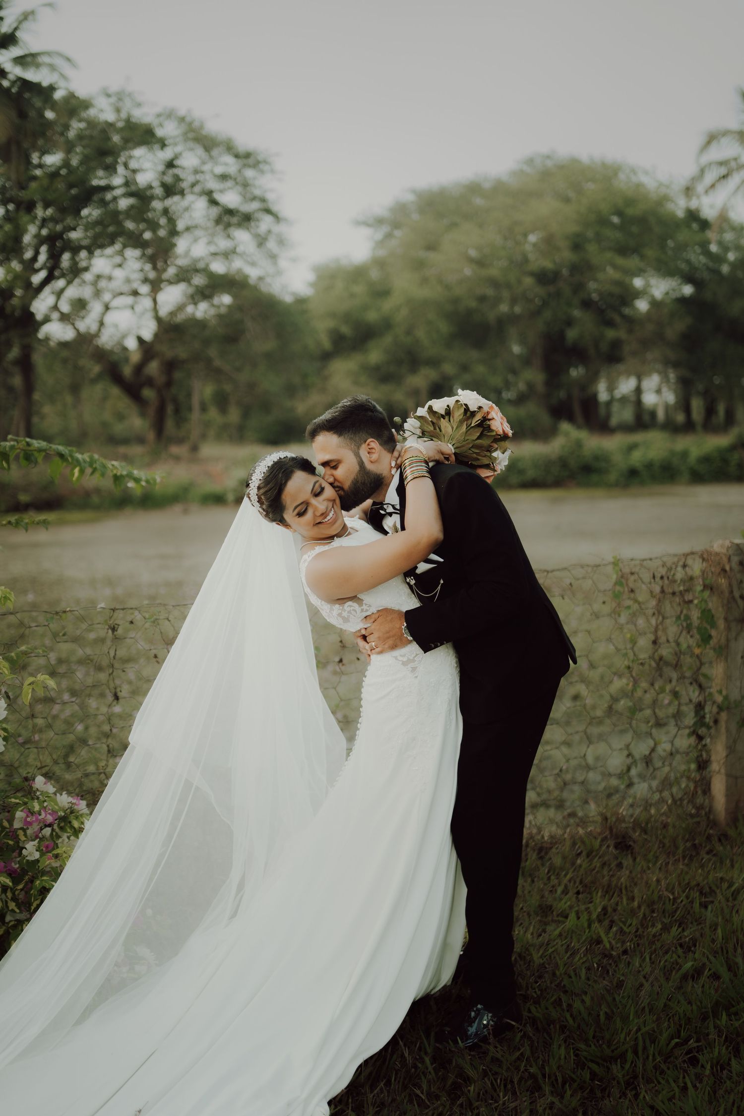 A bride and groom share a romantic embrace in a lush garden setting with a flowing white veil.