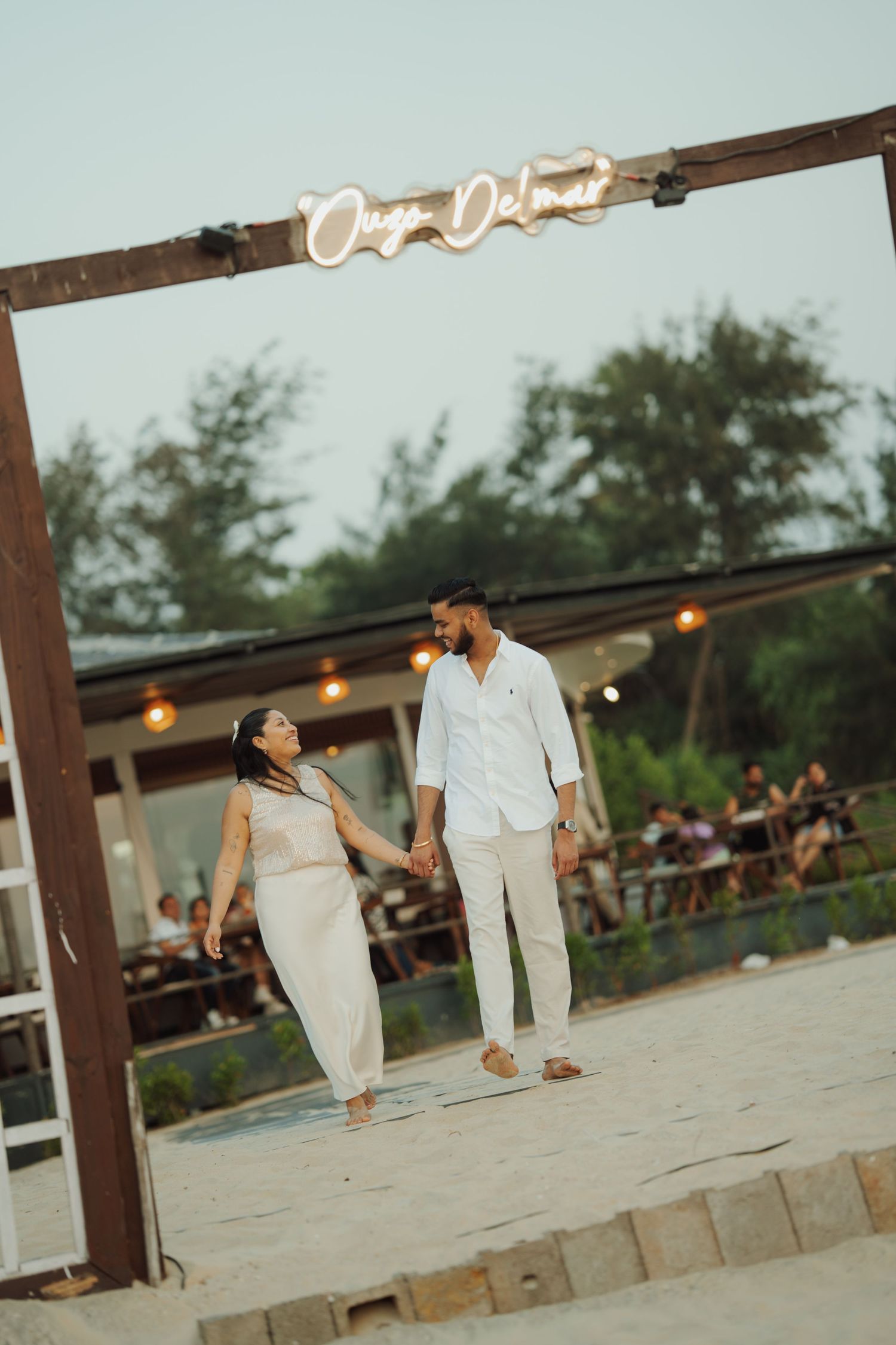 Couple holding hands under a wooden arch with neon sign on a sandy beach at dusk, dressed in elegant white outfits.