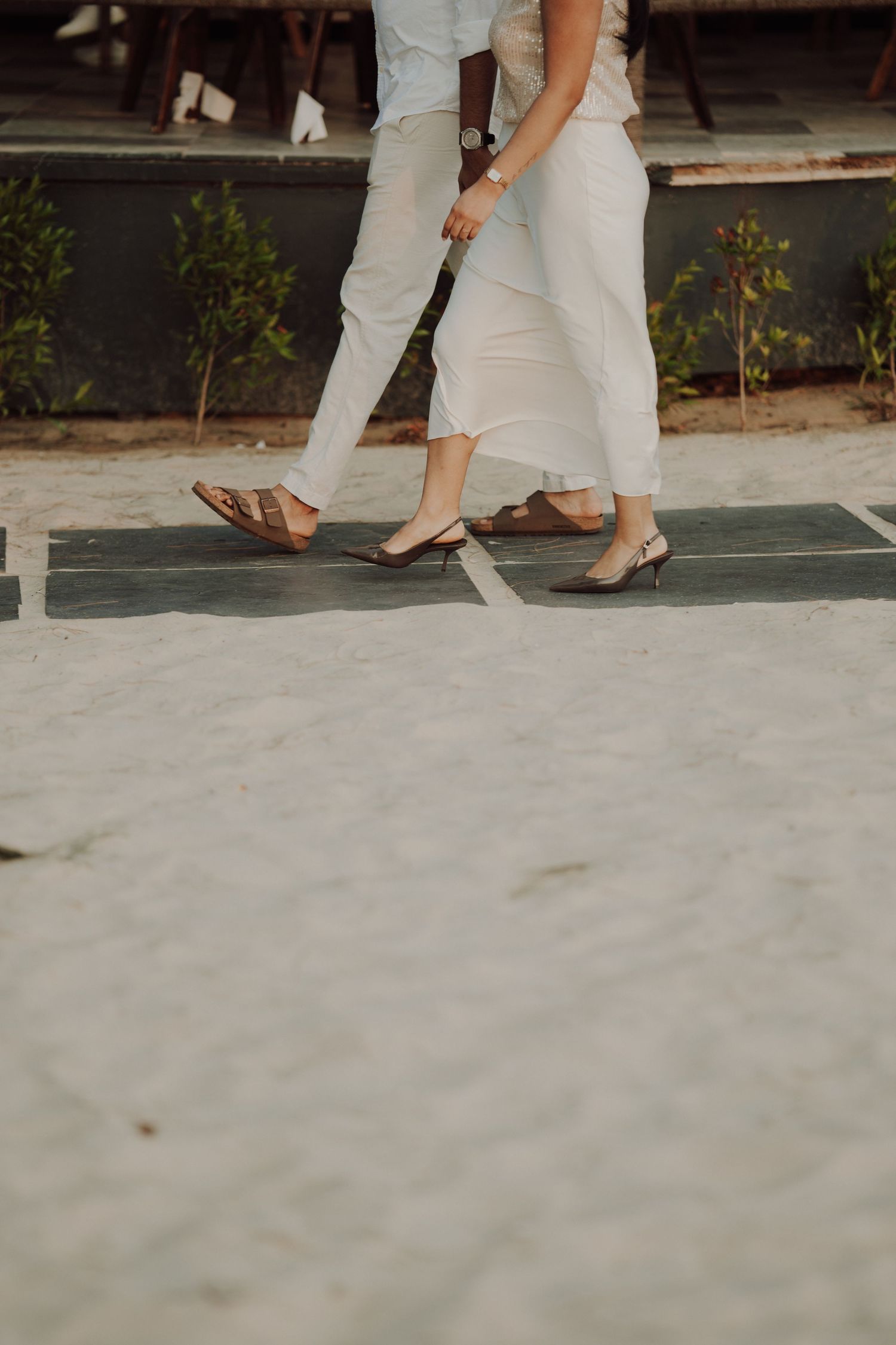 Couple walking together in white outfits, man in sandals and woman in heels, on a sandy path with greenery.